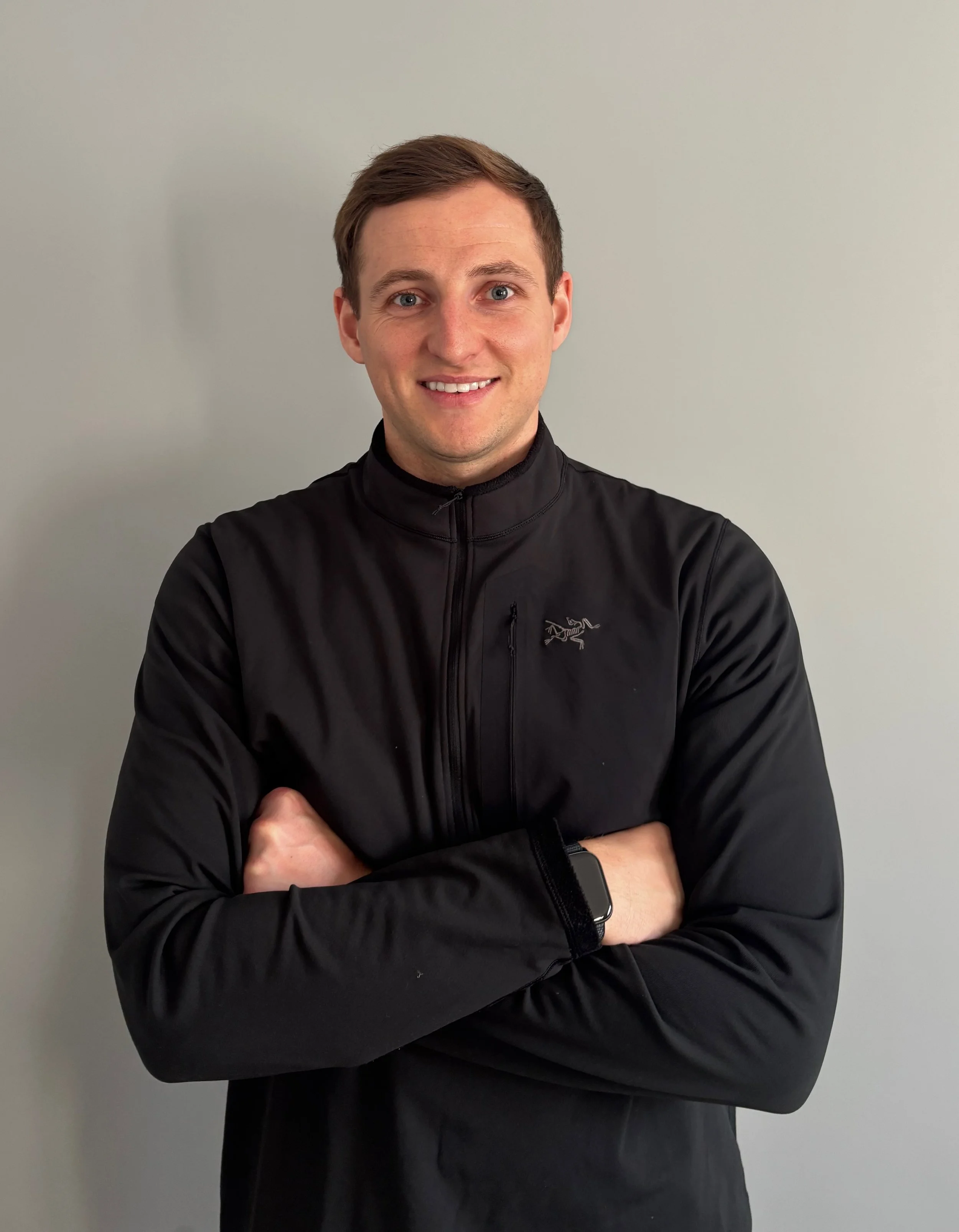 A young man with brown hair, smiling, standing against a plain light gray wall, wearing a black zip-up jacket with an animal logo and a smartwatch on his left wrist.