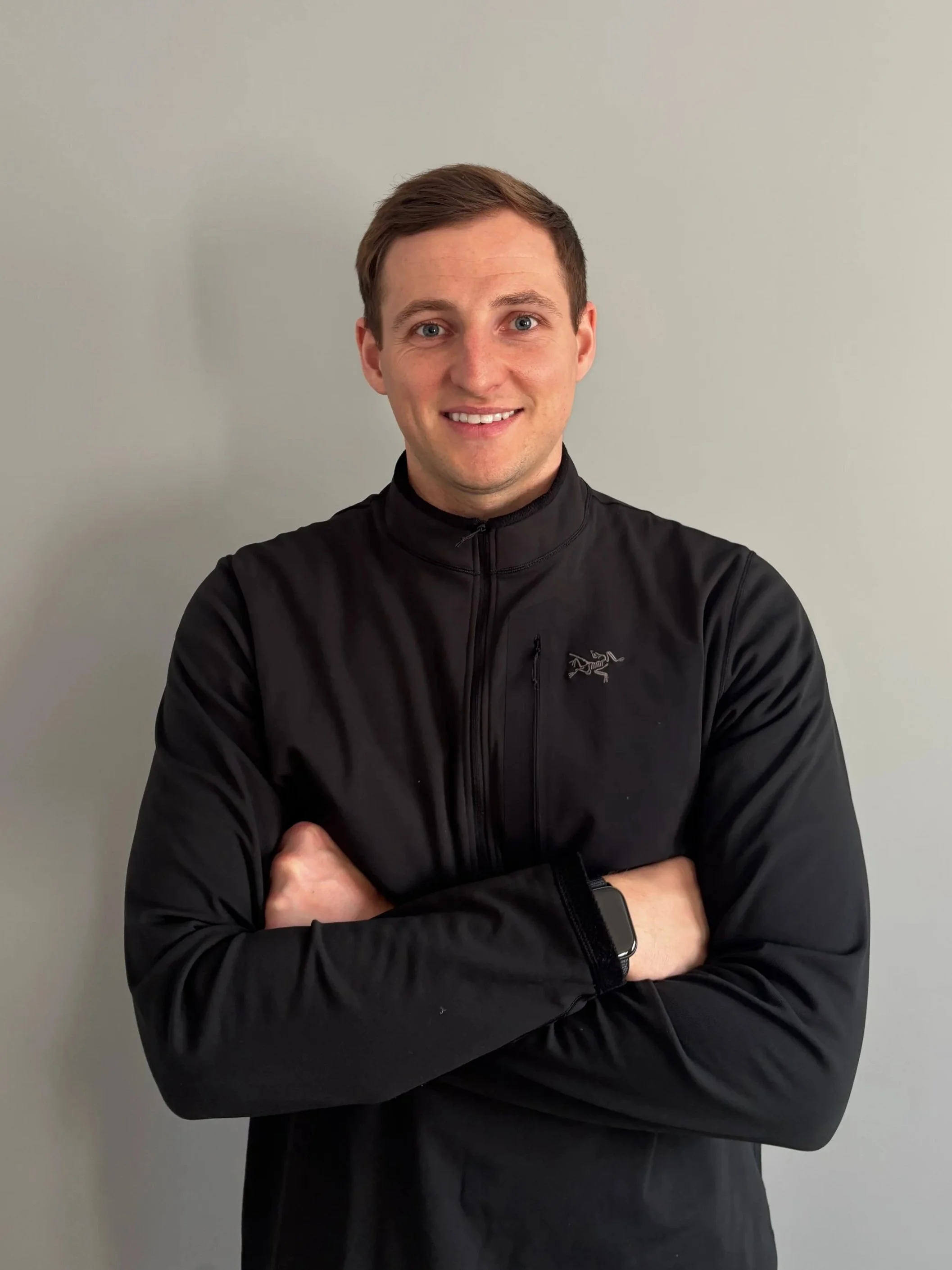 A young man with brown hair and blue eyes wearing a black jacket and smartwatch, standing with arms crossed in front of a plain light gray wall.