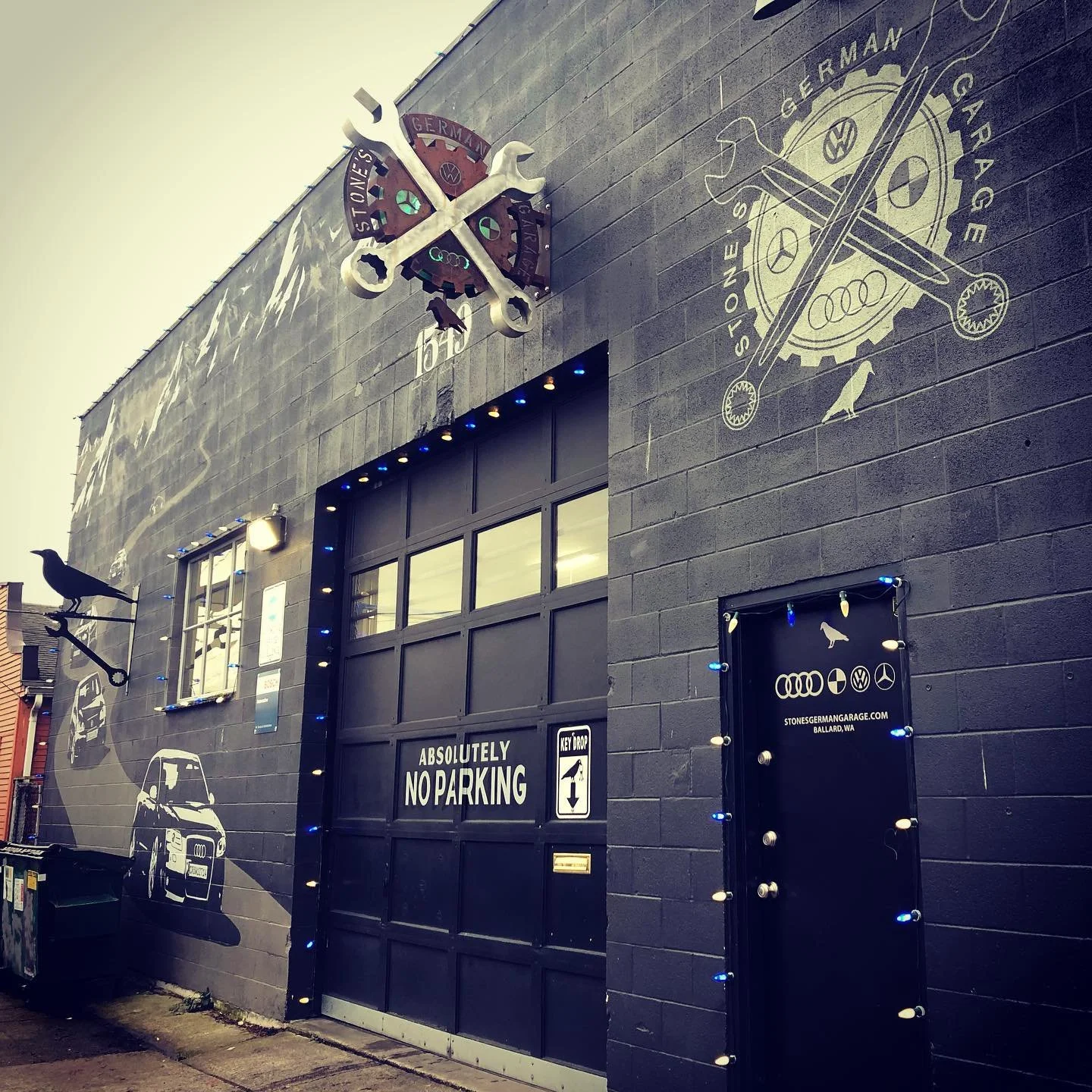 Exterior view of a garage with automotive-themed murals, including a silhouette of an Audi car and gear and tool graphics, decorated with blue holiday lights, and featuring a sign that reads 'Absolutely No Parking'.