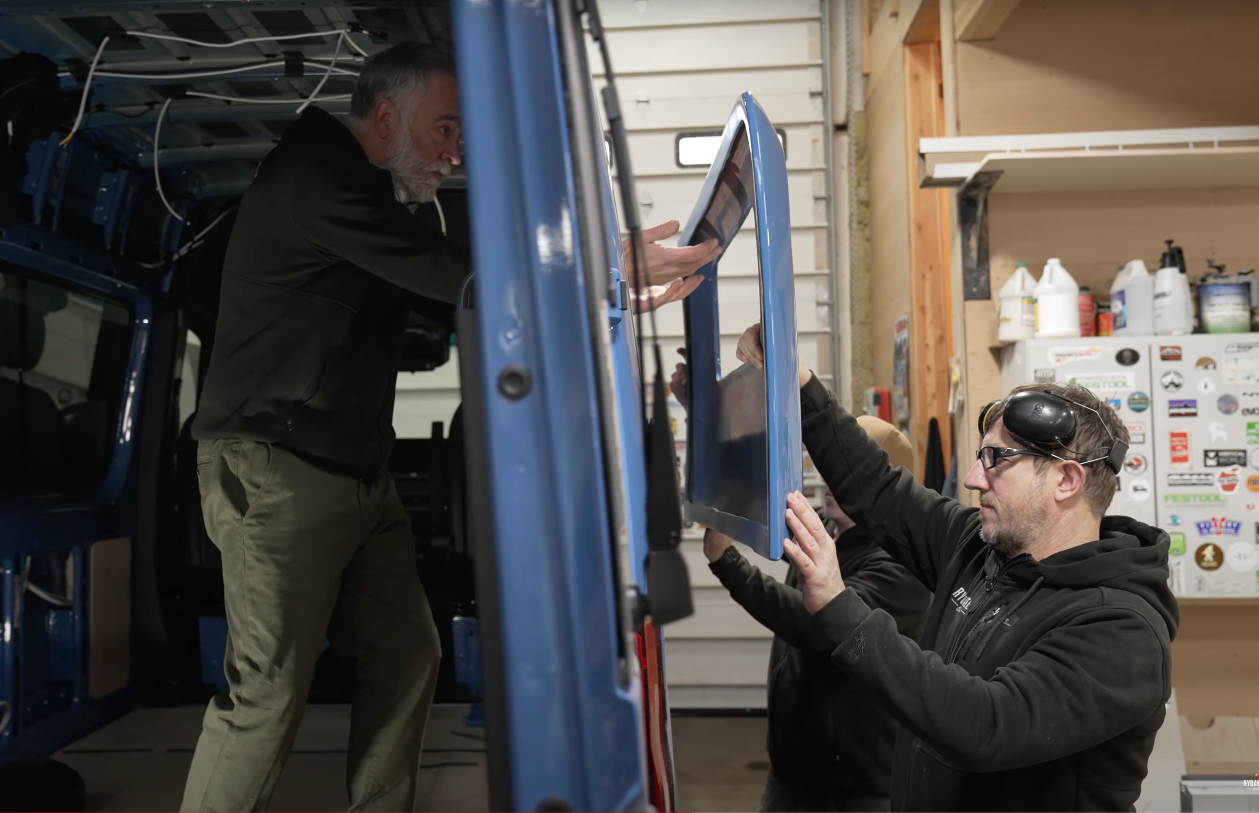Two men working on a blue vehicle in a garage, one standing and using a tablet, the other kneeling and handling a screen.