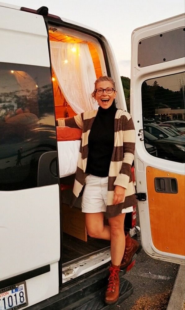 A woman smiling and posing in the entrance of a camper van with a wooden and white interior, during sunset in a parking lot.