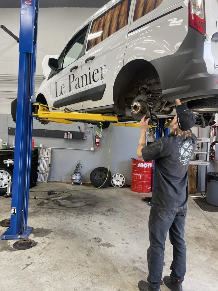 A mechanic working underneath a white van lifted on a hydraulic lift in an auto repair shop.