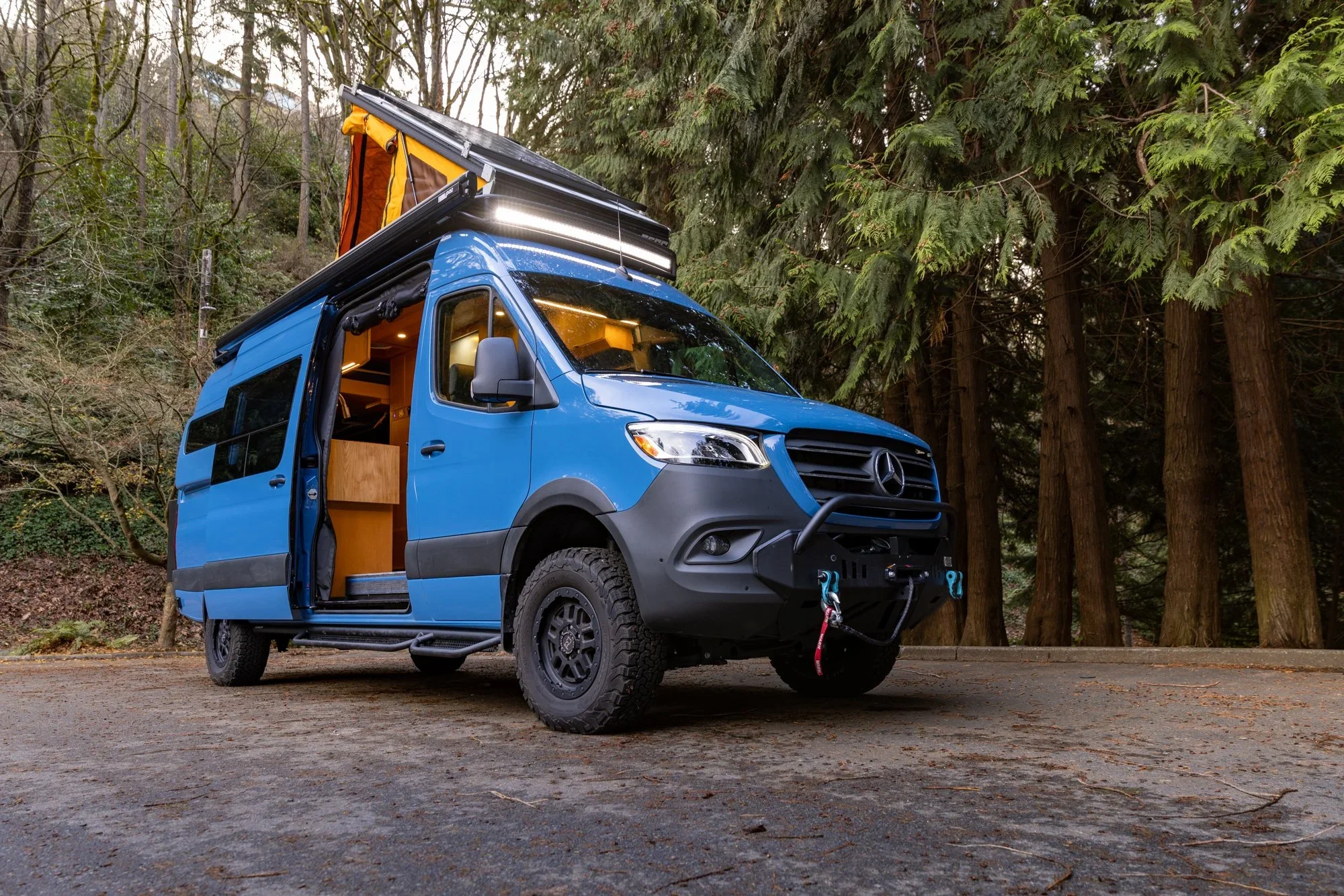 A blue camper van equipped for camping, parked in a forested area with tall trees in the background, and part of the van's interior visible through an open sliding door.