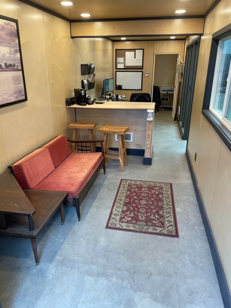 A small reception area with a pink velvet sofa, a wooden side table, a rustic wooden counter with two stools, a computer, and office supplies. There is a window on the right and a framed landscape photo on the left wall. The floor is concrete, and a 