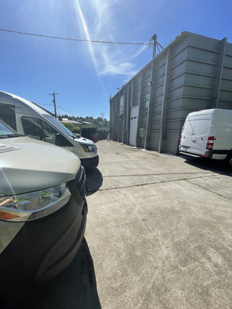 Parking lot with several parked vehicles, including a white van, a white SUV, and another white van, outside a gray industrial building under a bright sunny sky.