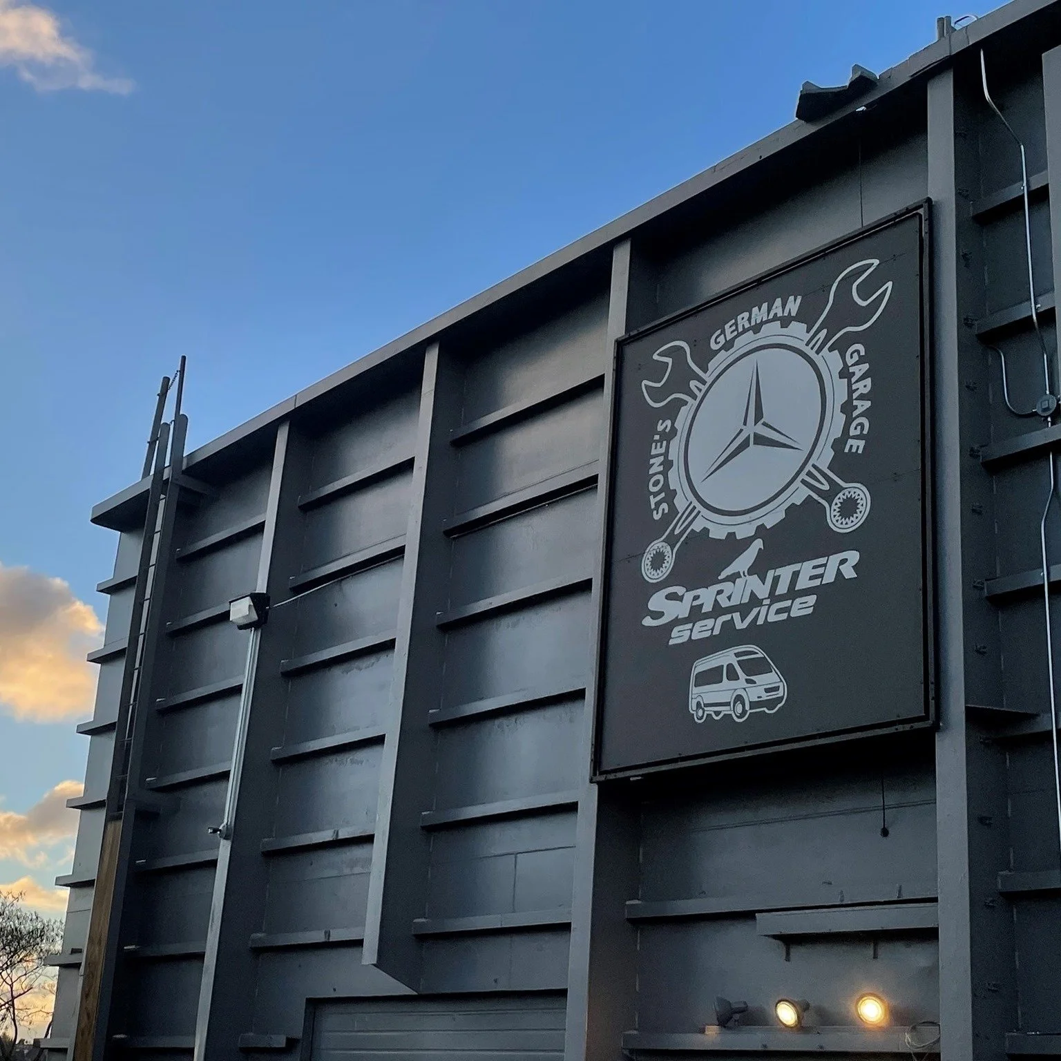 Building with a sign for Stones German Garage, offering Sprinter service, featuring a logo with a gear, wrenches, and a Mercedes-Benz emblem.