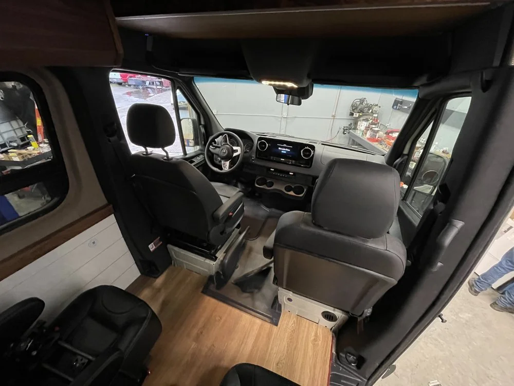 Interior view of a camper van with two front seats and a digital dashboard, inside a garage or workshop.