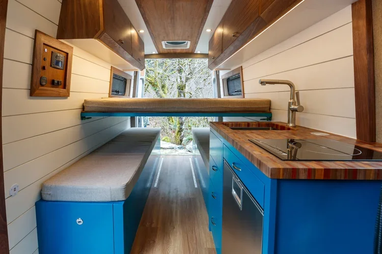 Interior of a tiny home or camper with blue cabinetry, a small countertop with a stovetop, a sink with a modern faucet, a seating bench, and a bed above the bench, with large windows showing trees outside.