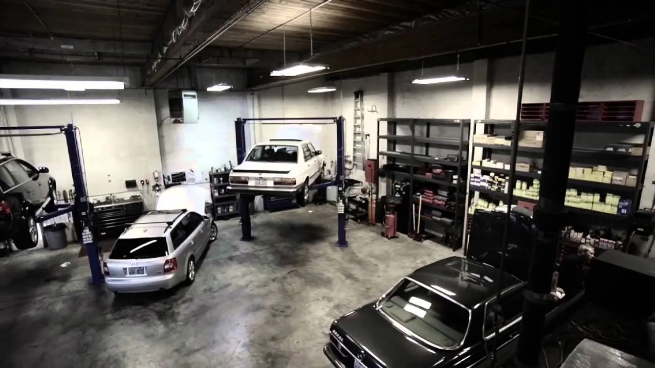 Auto repair garage with four cars, including one elevated on a hydraulic lift, and shelves filled with parts and tools.