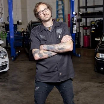 A man with glasses and tattoos stands with arms crossed in a garage or workshop surrounded by cars and equipment.
