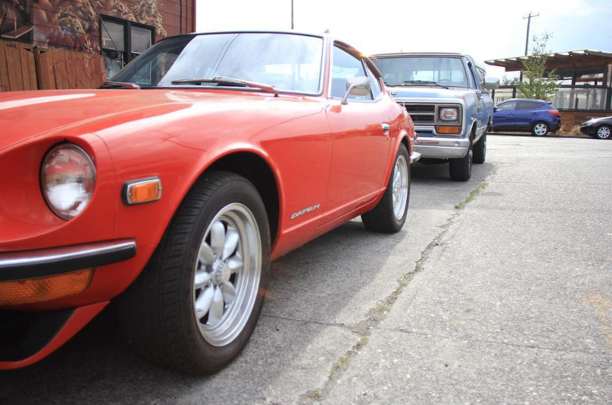 A red vintage Datsun sports car parked in a parking lot with other older and modern vehicles.
