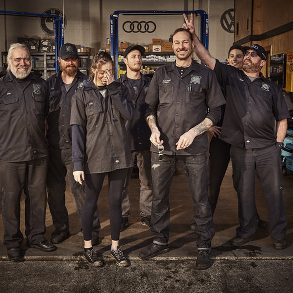 Group of six people wearing black mechanic uniforms standing in a garage with tools and equipment, one person making bunny ears behind another's head, smiling and posing.
