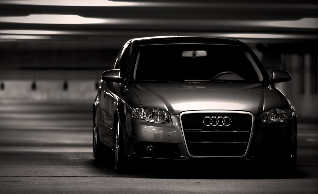 A dark gray Audi sedan in a parking garage or tunnel, illuminated by overhead lights, with a focus on the front of the car.