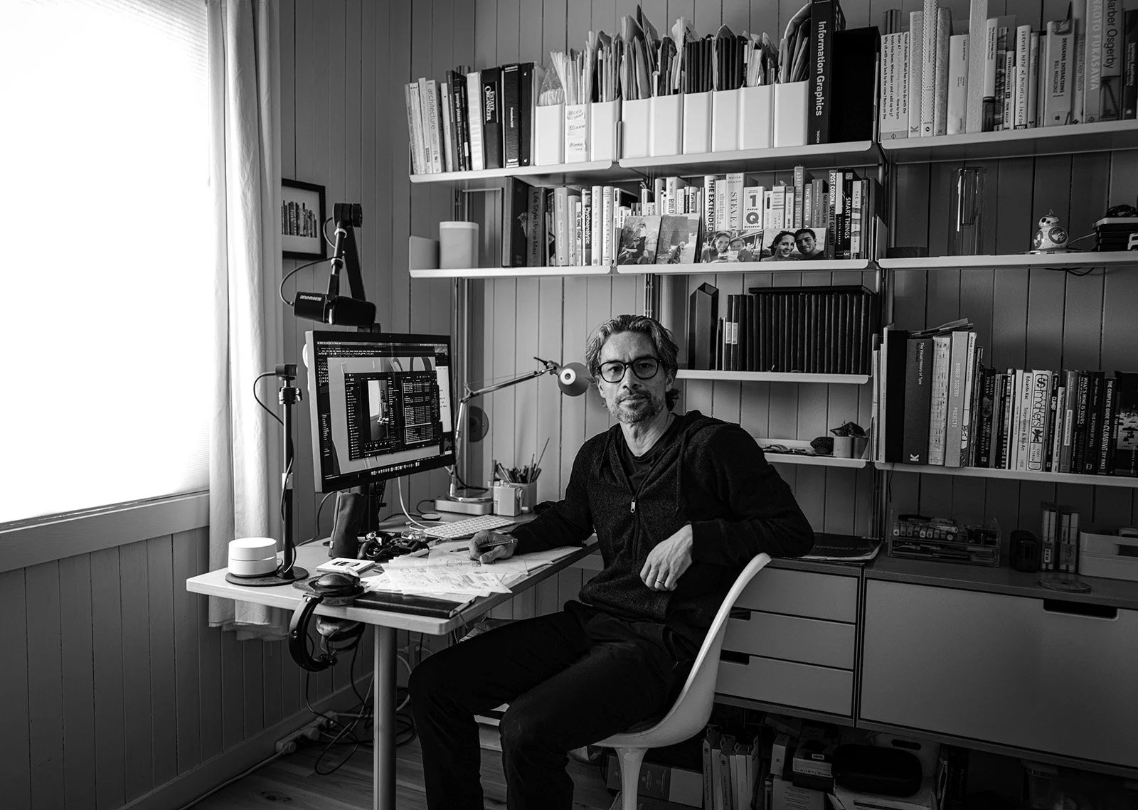 A man with glasses seated at a desk in a home office, with a computer monitor, bookshelves filled with books, and various office supplies.