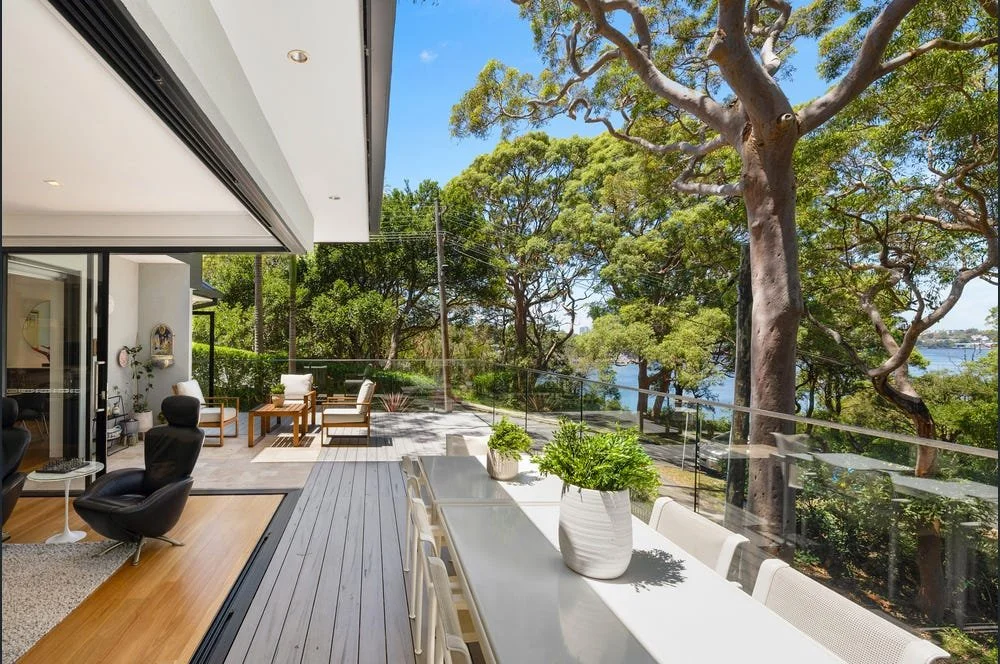Modern outdoor balcony with dining table, potted plants, and seating area overlooking lush green trees and Sydney Harbour