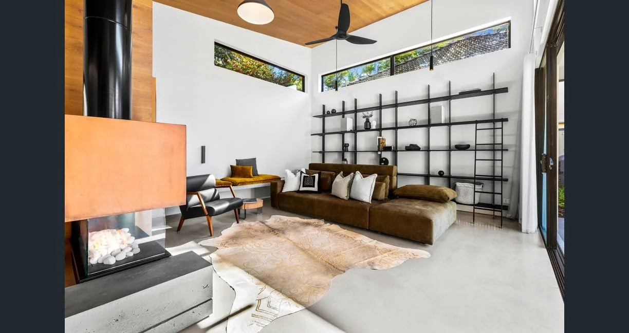 Modern living room with white walls, high ceiling, large window and black ceiling fan. Brown sectional sofa with pillows, glass shelving unit, black leather chair, beige rug, and decorative items.