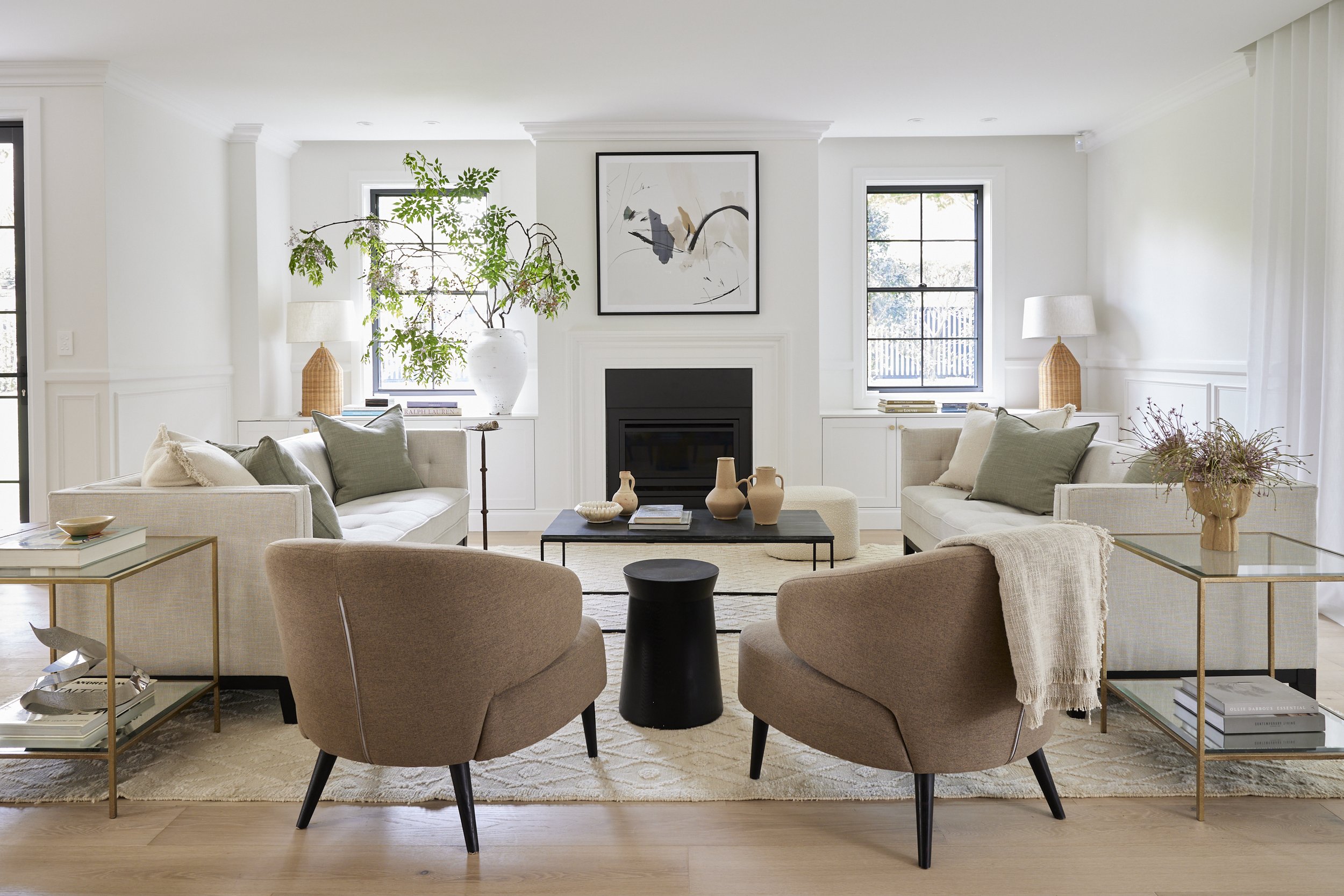 A bright, modern living room with white walls, a fireplace, large windows, and cozy seating including two beige armchairs and a sofa with pillows. Decor includes artwork above the fireplace, a large plant, lamps, books, and decorative vases, all arranged on glass and metal side tables.