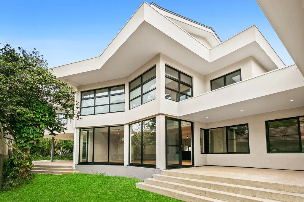 Modern white house with large windows and a multi-tiered roof, surrounded by green grass and trees.