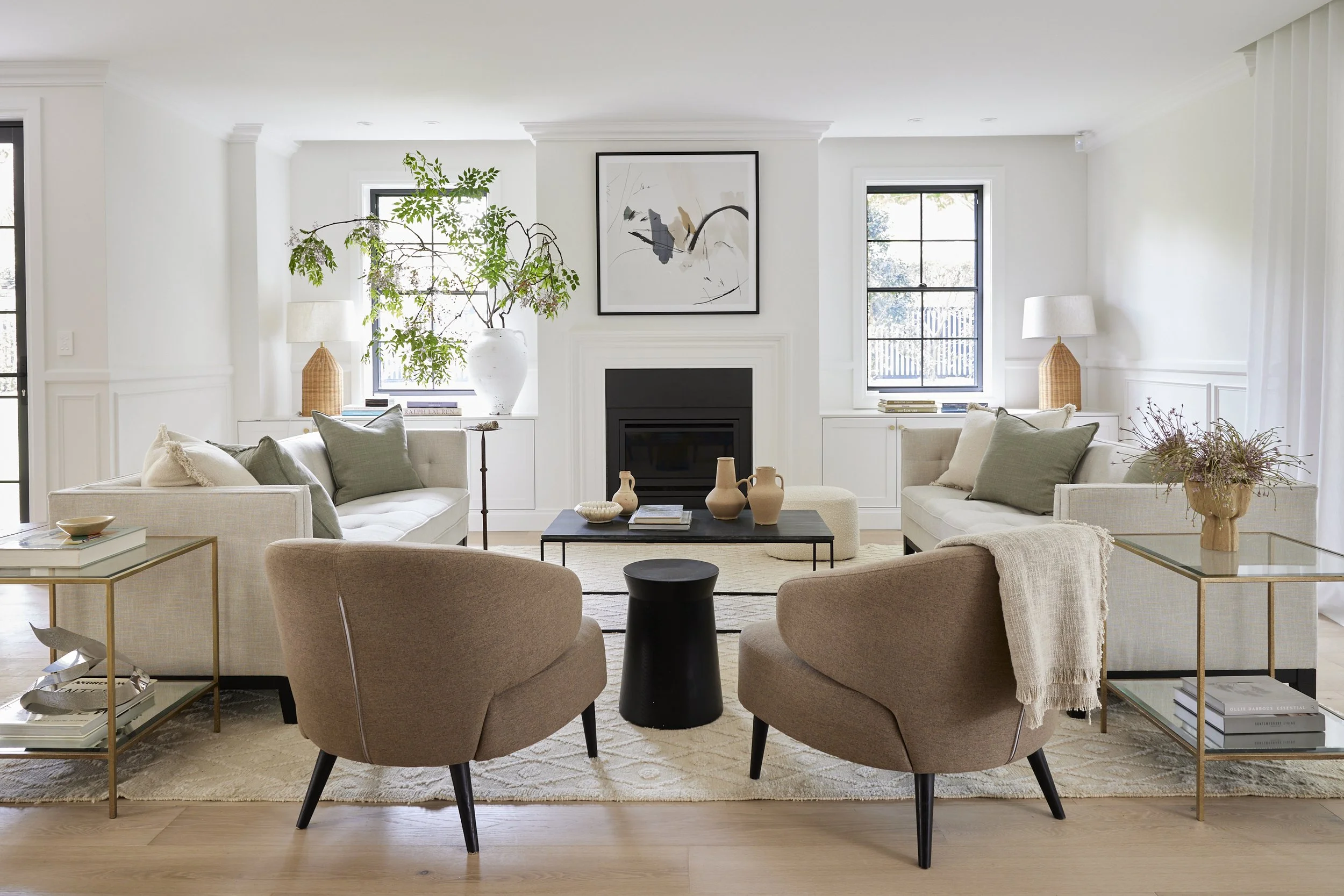 Bright modern living room with white walls and ceiling, featuring a black fireplace, abstract framed art, large windows, beige couches with pillows, a black side table, decorative vases, lamps, and indoor plants.
