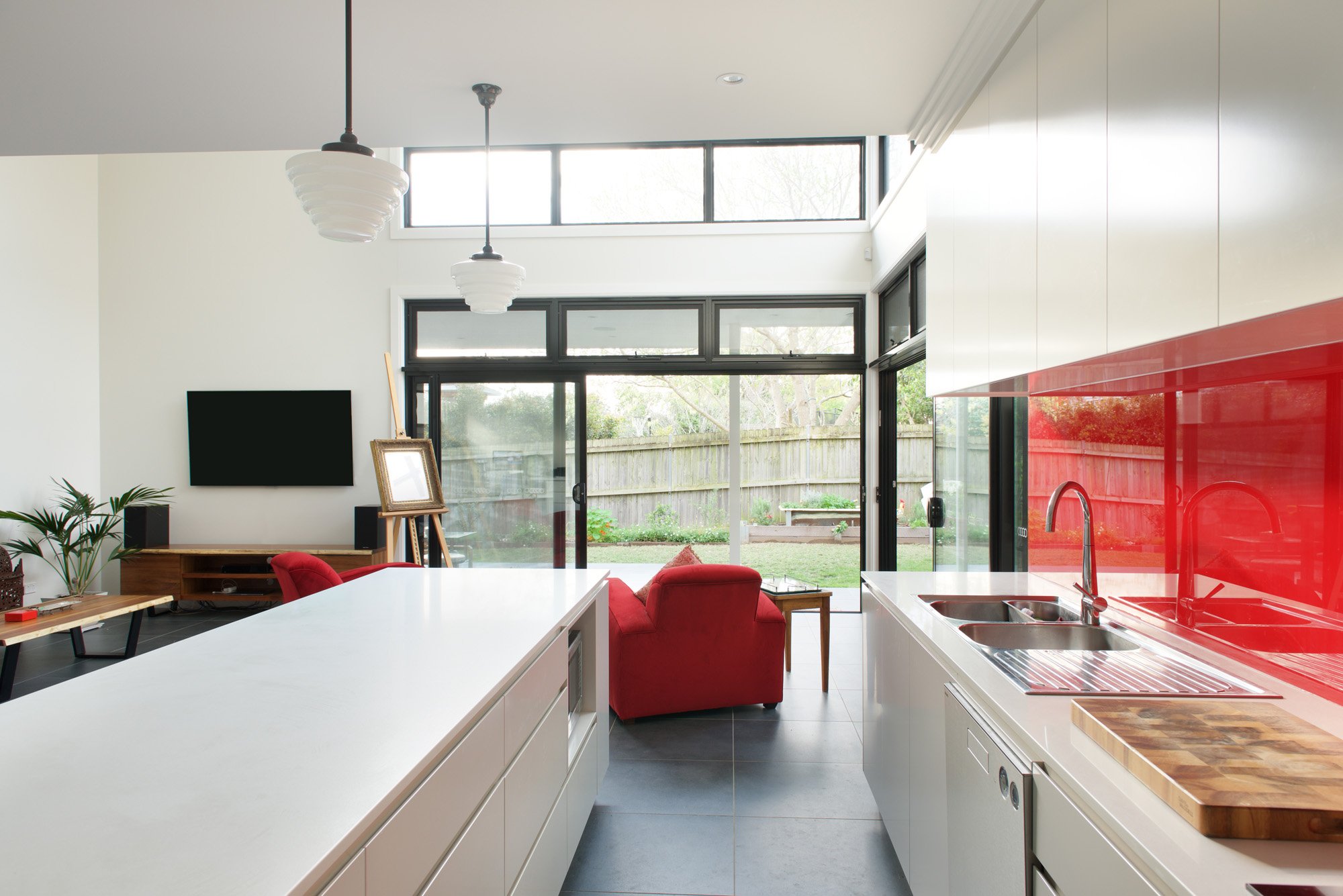 A modern, open-concept kitchen and living space with large sliding glass doors leading to a backyard. The kitchen has a white island with a wooden cutting board on top, and white cabinetry with a red backsplash. There are two white pendant lights hanging from the ceiling. The living area includes a red sofa facing a wall-mounted flat-screen TV, situated beneath a set of high windows that allow natural light. There are plants on a wooden table and other decor elements in the space.