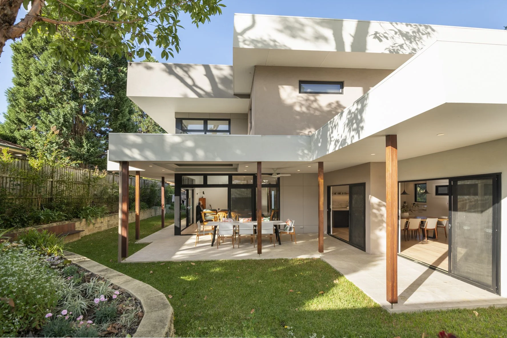 Modern two-story house with an open patio, outdoor dining table with chairs, sliding glass doors, and a garden with plants and trees.