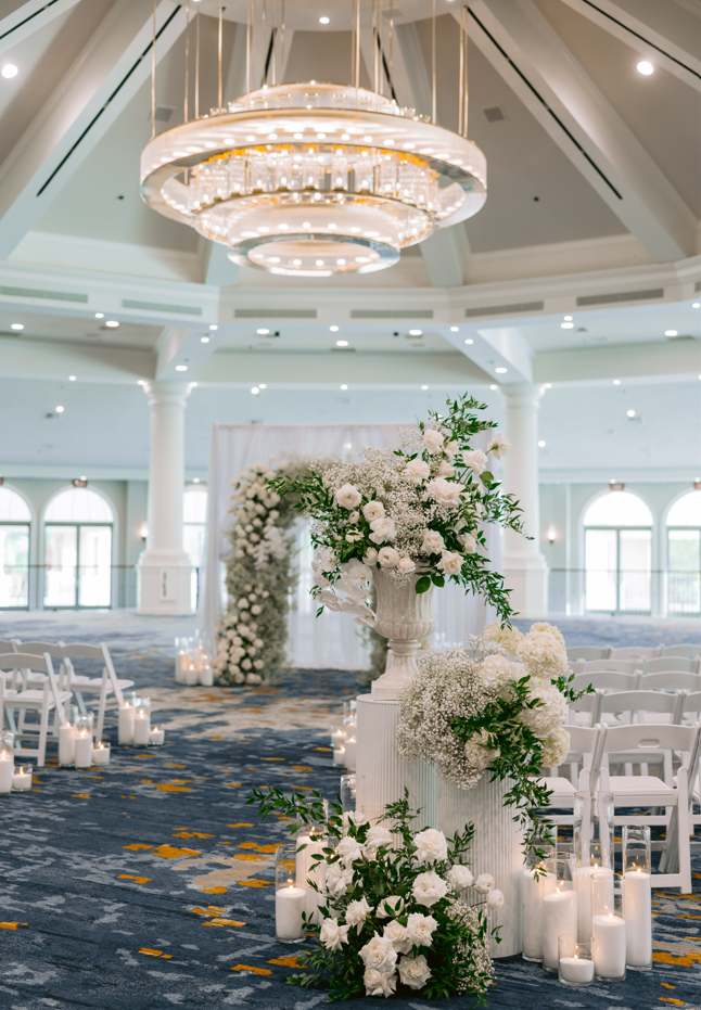 White floral arrangement with roses and baby's breath on a column inside a wedding ceremony hall, with candles on the floor and chairs arranged for guests.