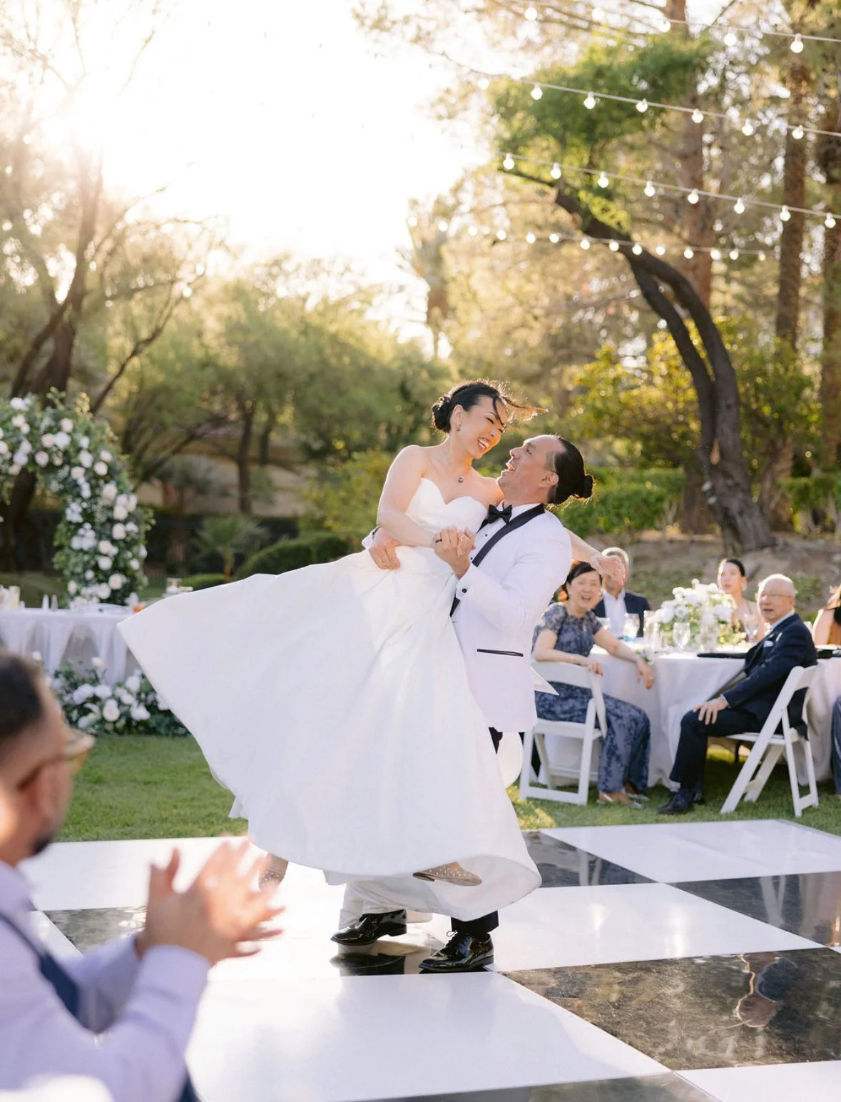 A bride and groom dancing at an outdoor wedding reception during sunset, with guests seated at tables in the background.