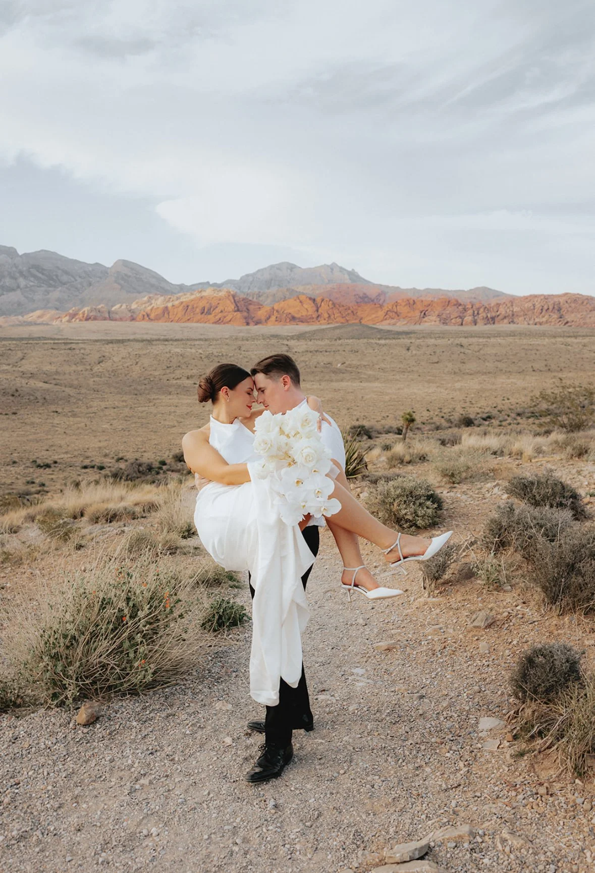 A couple in wedding attire in a desert landscape with mountains in the background. The man carries the woman in his arms, and they are touching foreheads. The woman is holding a large bouquet of white flowers, and both are dressed in white, with the 