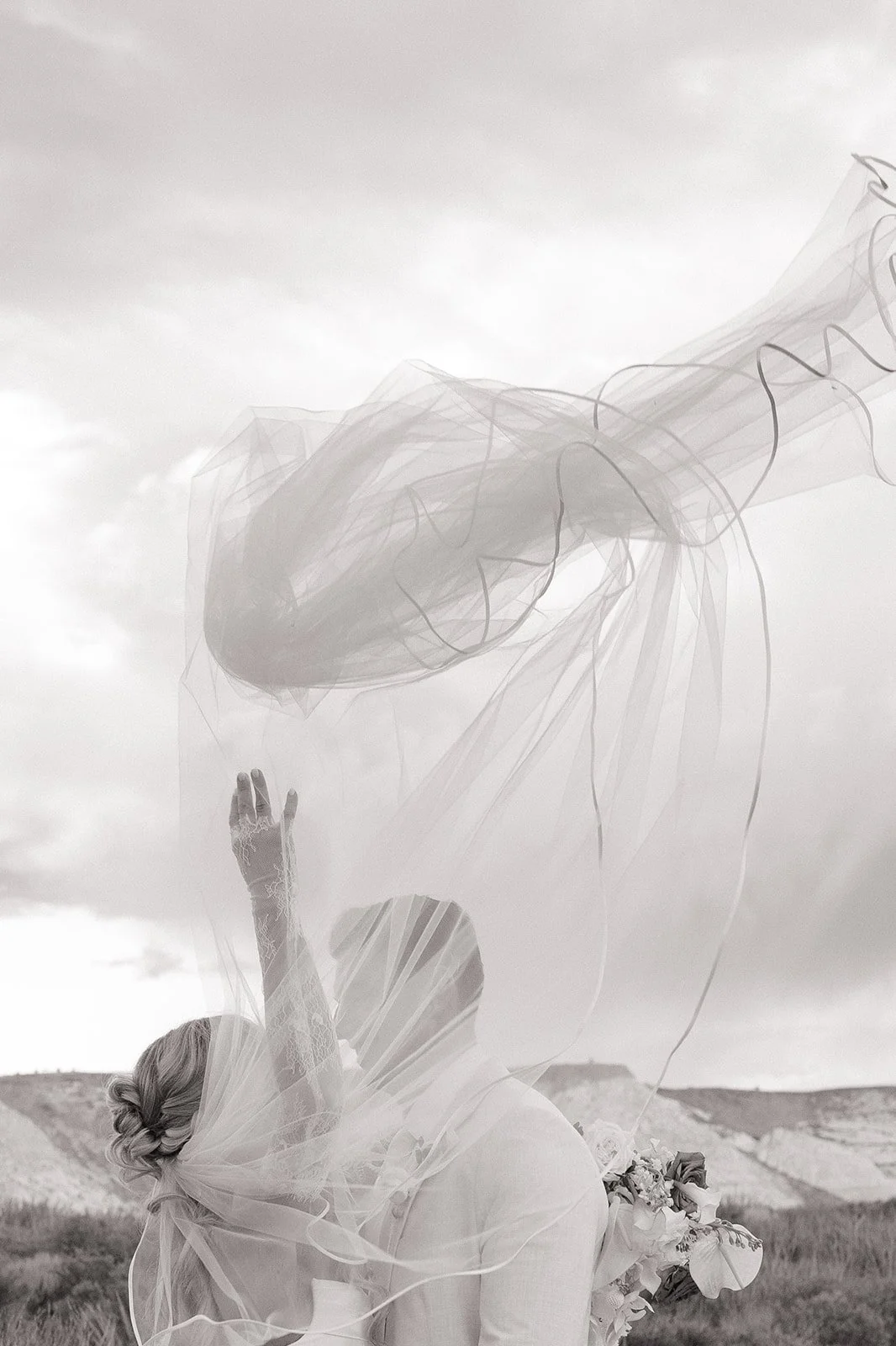 A bride holding a bouquet with her head tilted back, raising one hand, and a large, translucent tulle veil and ribbon-like fabric swirling above her against a cloudy sky.