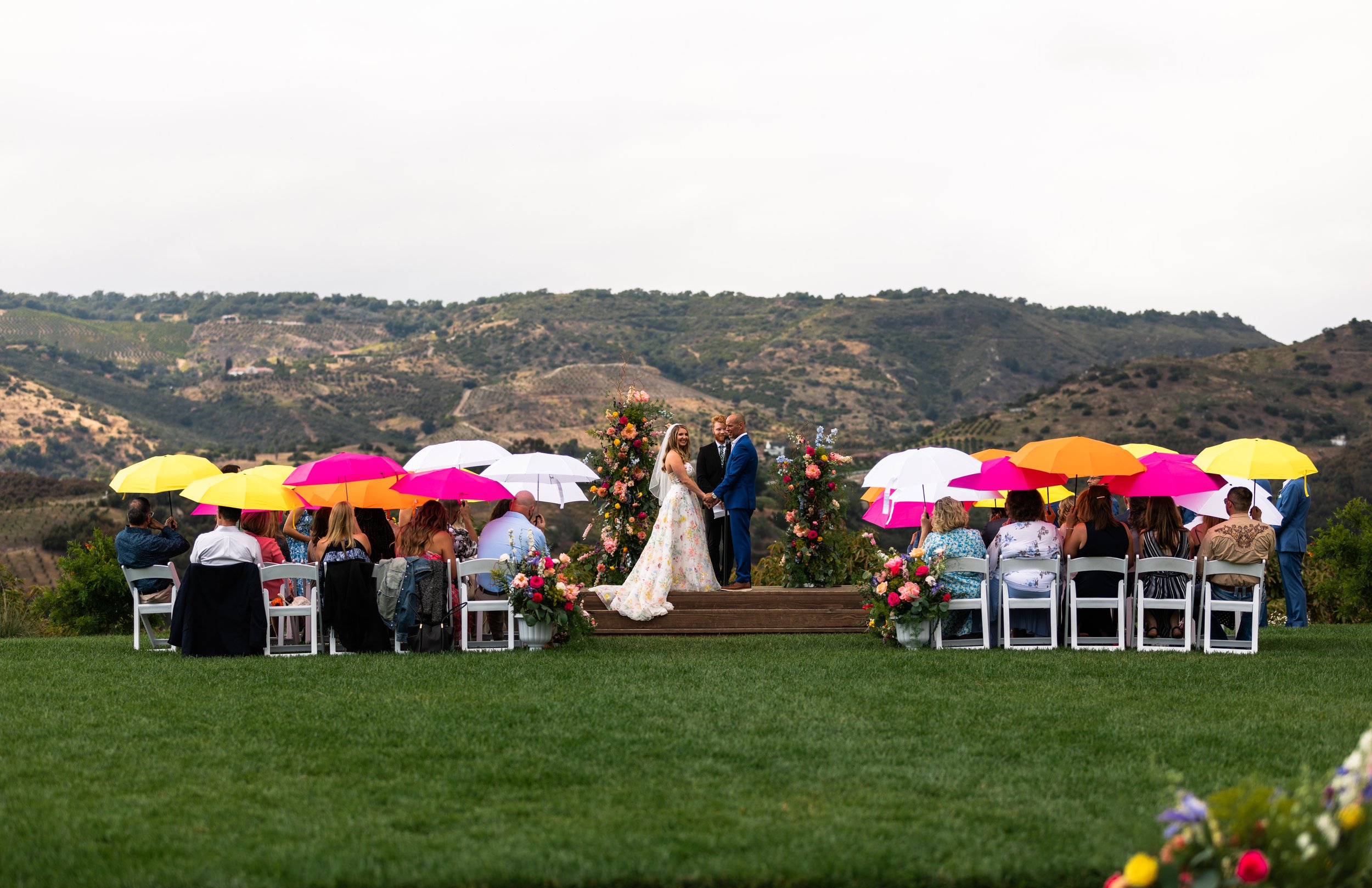 A wedding ceremony outdoors with a bride in a floral gown and groom in a blue suit, standing on a wooden platform decorated with flowers. Guests are seated under colorful umbrellas on a grassy area, with rolling hills in the background.