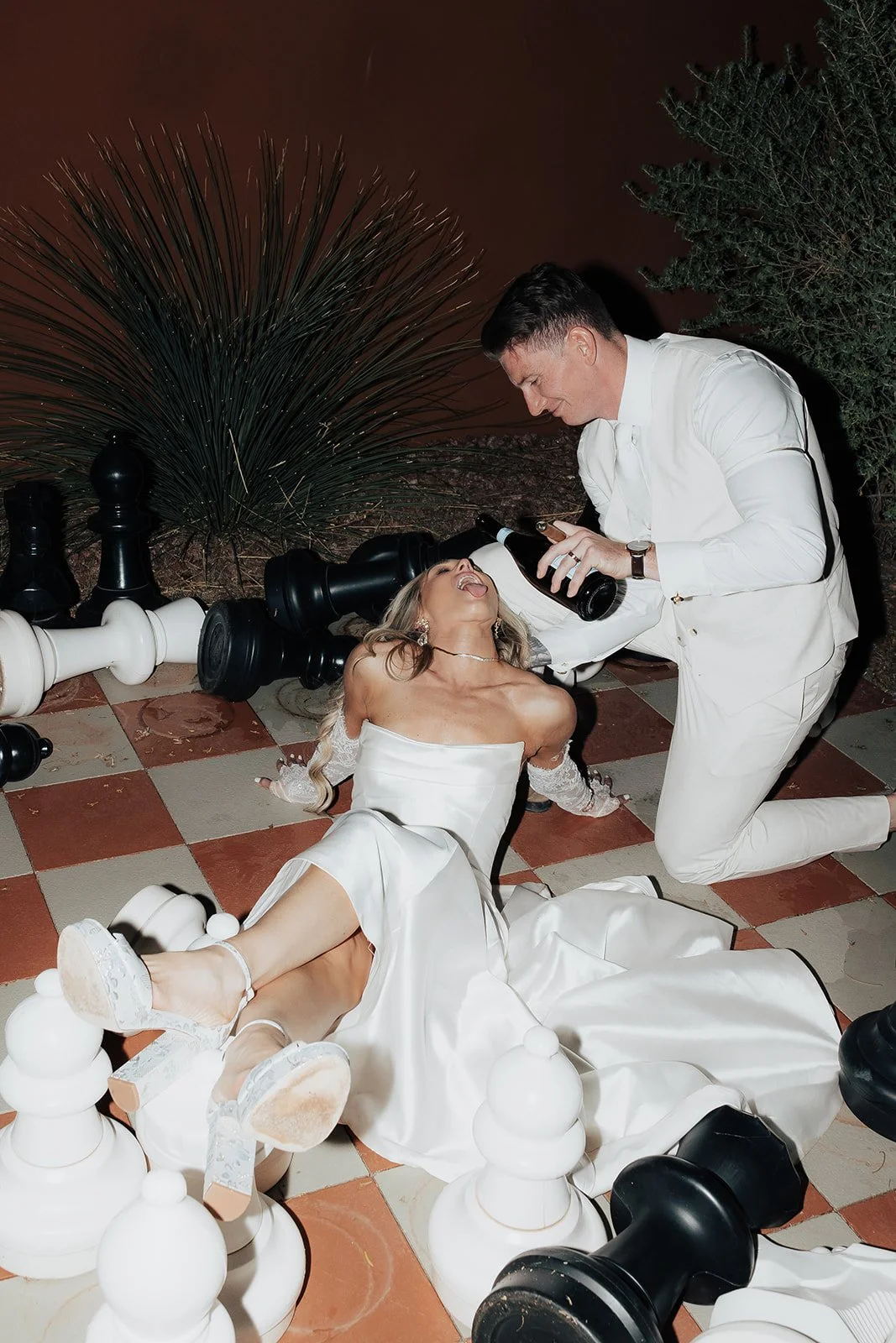 A woman in a wedding dress is on the ground surrounded by toppled large chess pieces. A man in white kneels beside her, holding a bottle and pouring liquid onto her.