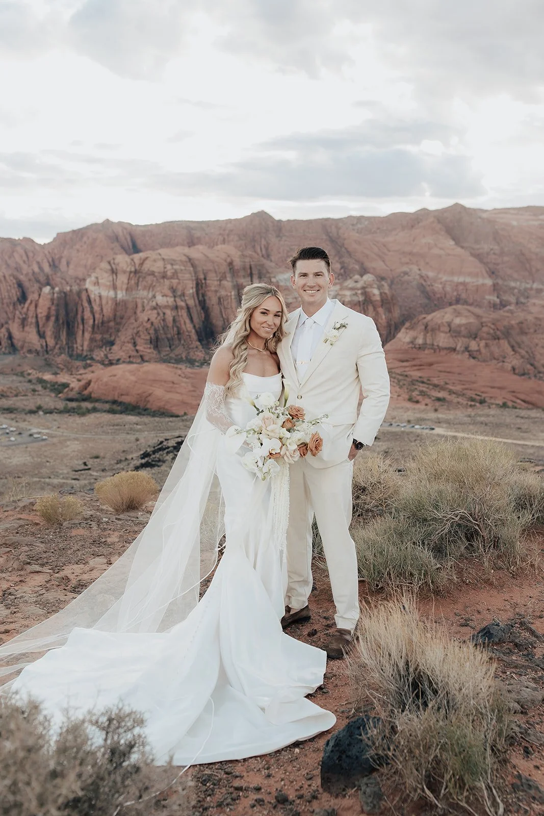 A bride and groom in wedding attire standing in a desert-like landscape with red rocks and mountains in the background, holding a bouquet of flowers.