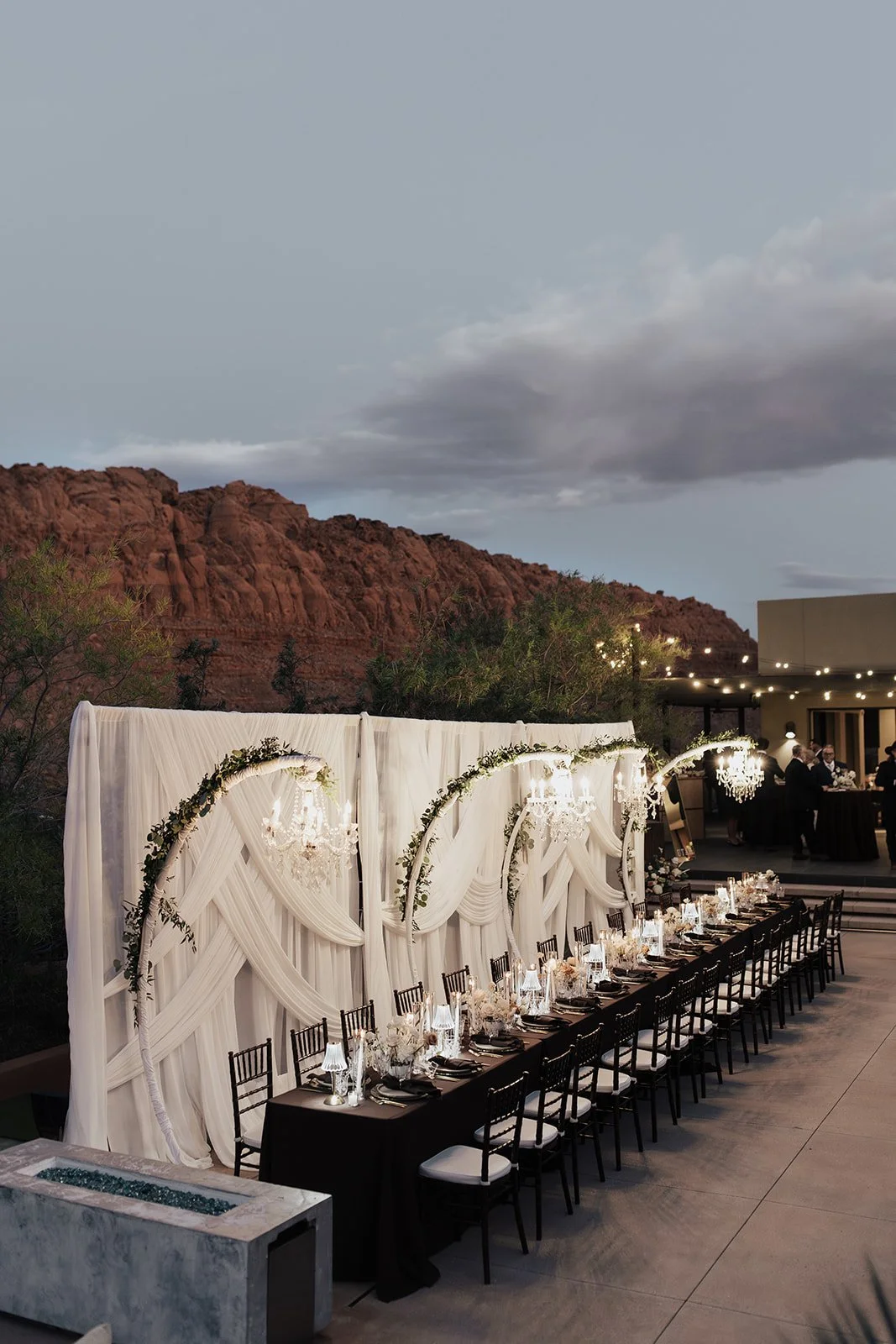 An outdoor wedding reception setup with a long black table, elegant white drapery backdrop, chandeliers, and string lights, set against a rocky mountain landscape in the evening.