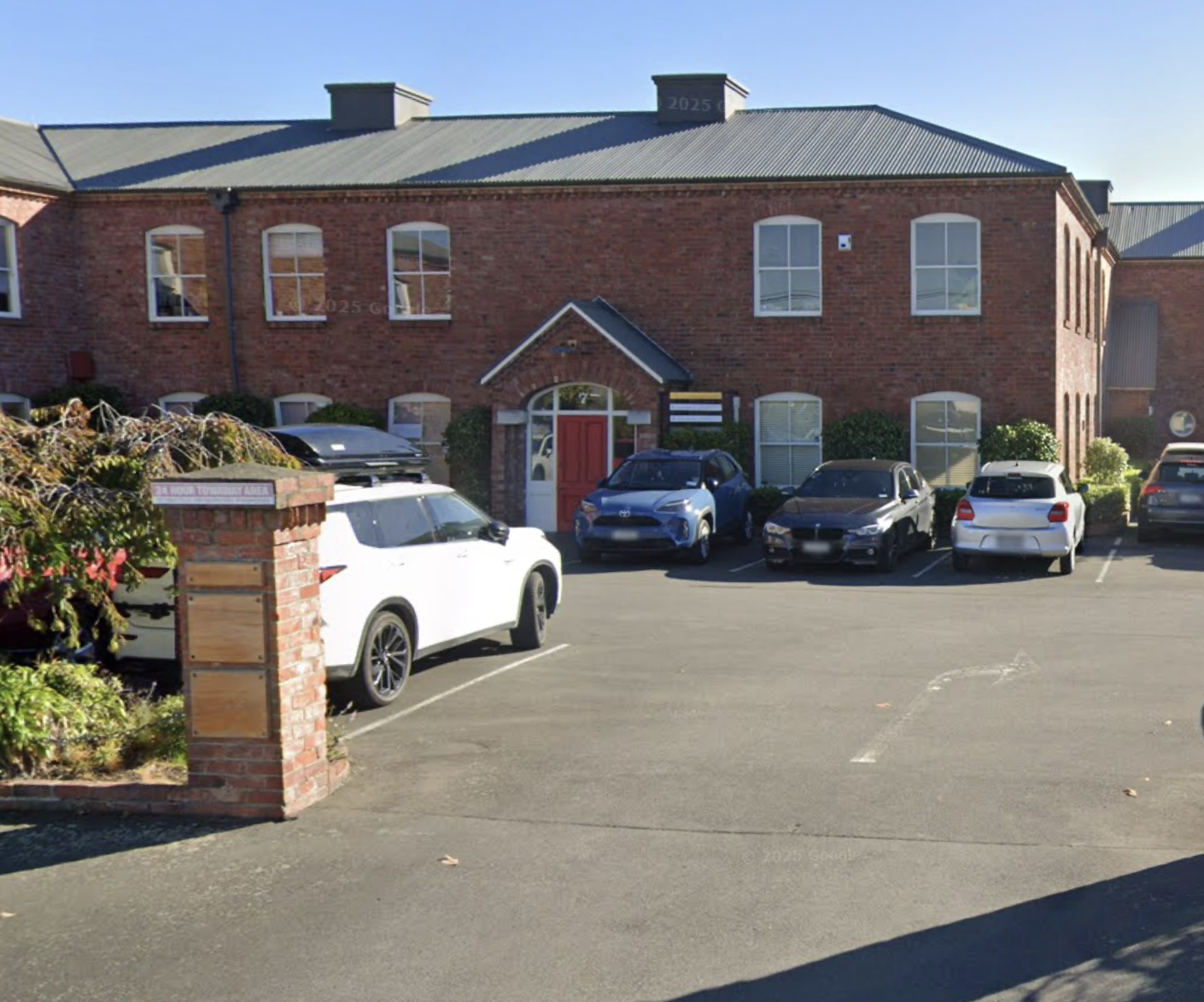 Parking lot in front of a red brick building with six cars parked and a brick wall with a small wooden section in the foreground.