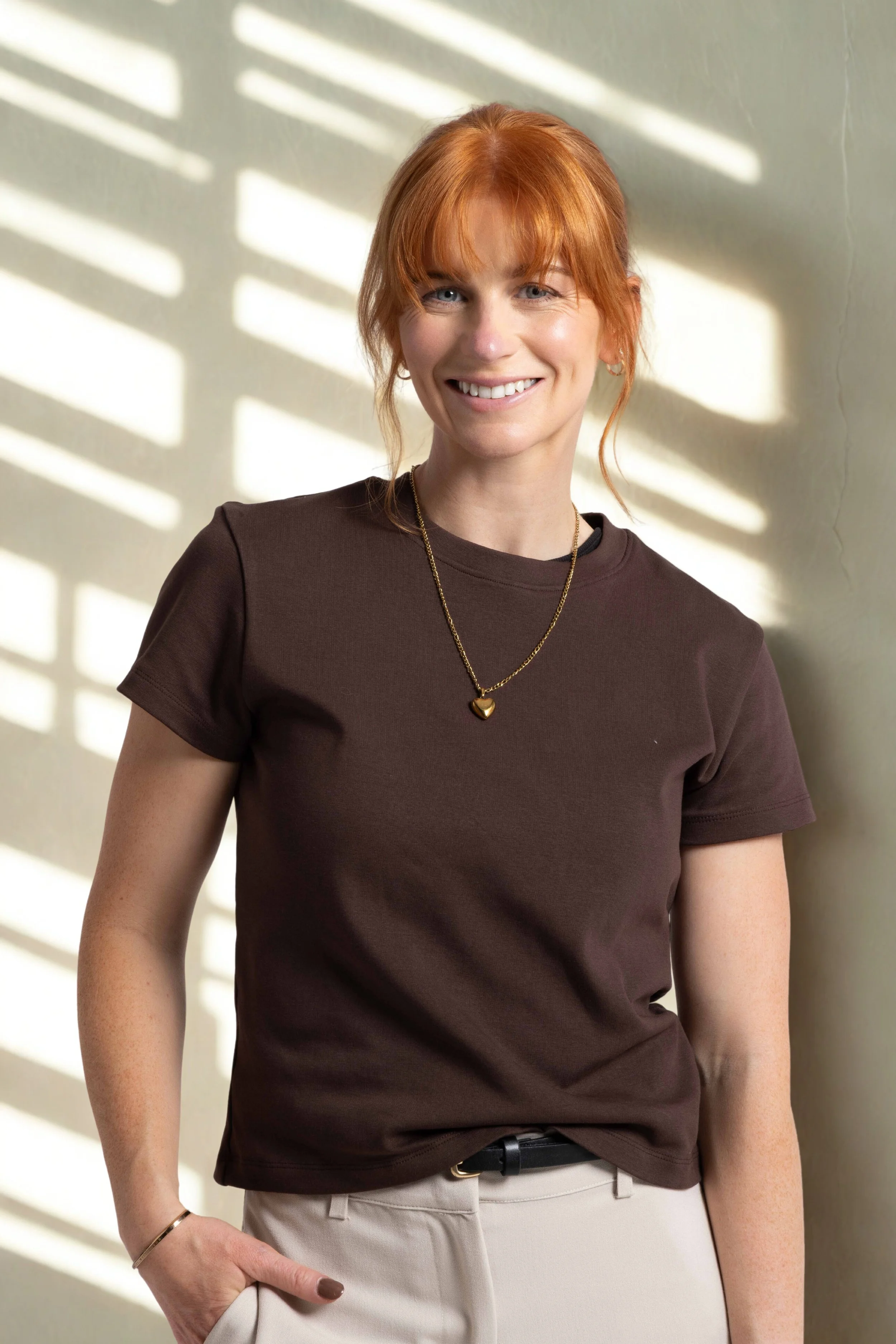 Woman with red hair wearing a brown T-shirt, beige pants, and gold jewelry standing against a wall with sunlight and shadow patterns.