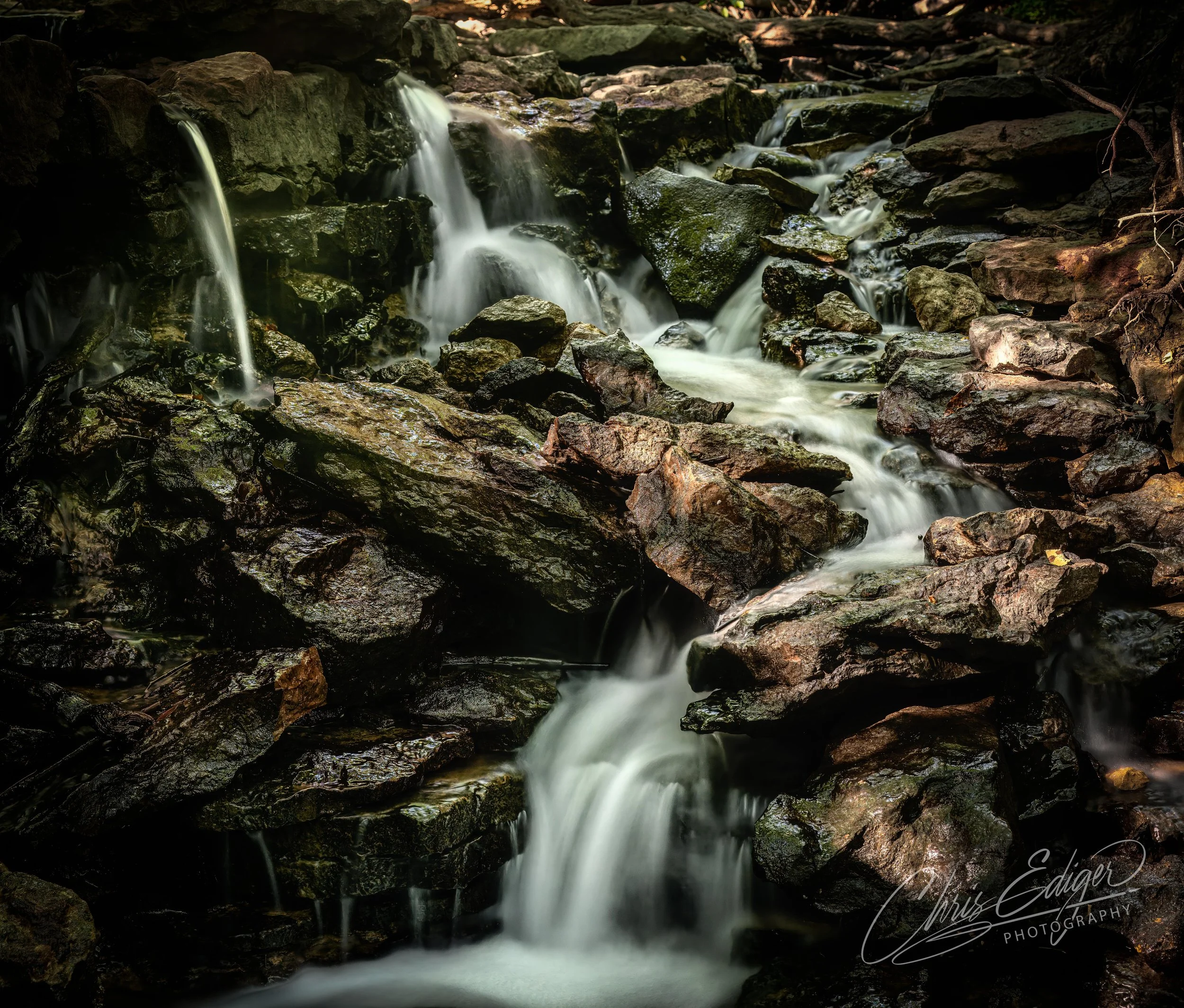 A long exposure photograph captures the ethereal, silky flow of water cascading over a dark, moss-covered jumble of rocks. The light filters through the canopy, illuminating the scene and highlighting the texture of the wet stones in this tranquil, n