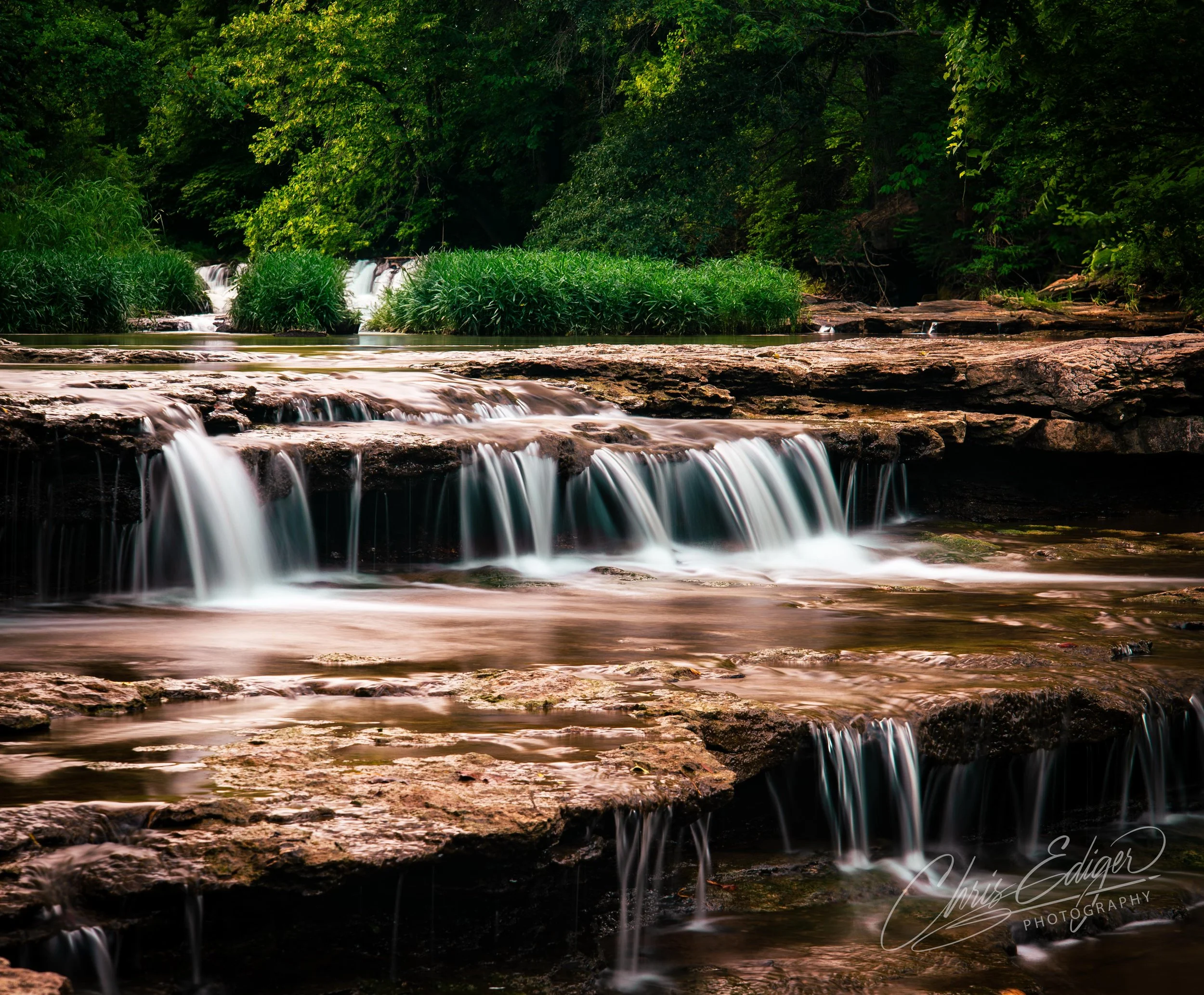 A multi-tiered waterfall flowing over rocks with green vegetation and trees in the background.