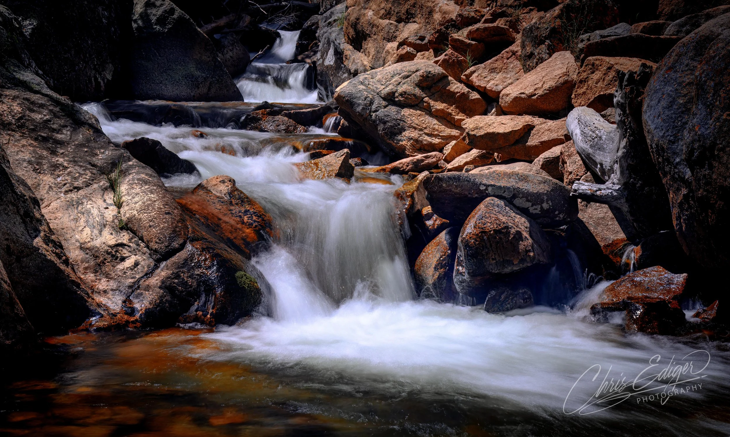 A serene long-exposure photograph of a small mountain stream tumbling over rugged rocks. The silky motion of the water is captured in smooth white flows as it winds its way through a natural stone staircase, framed by warm-toned boulders and earthy t