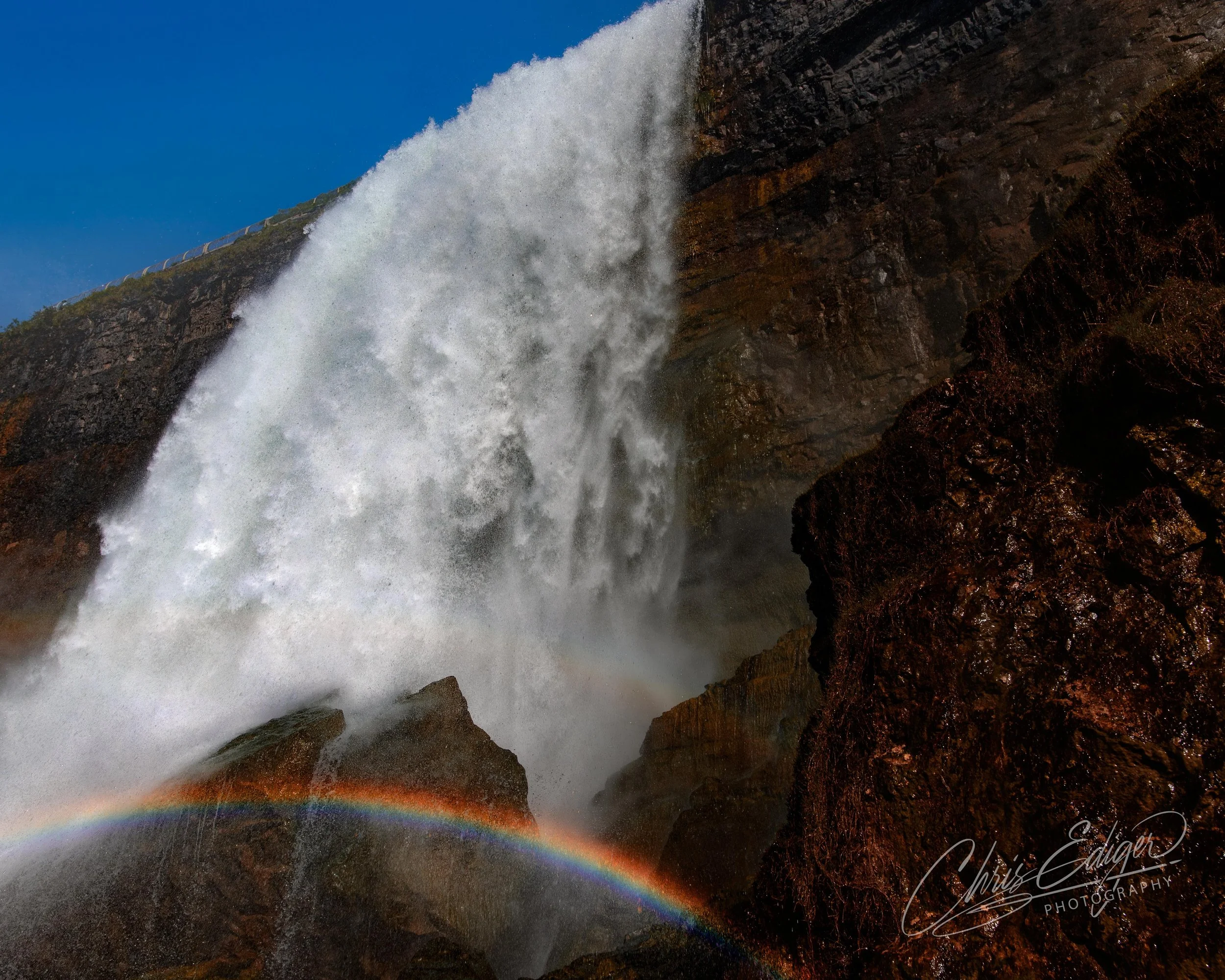 A large waterfall crashing down over rocks with a rainbow at the base in bright daylight.