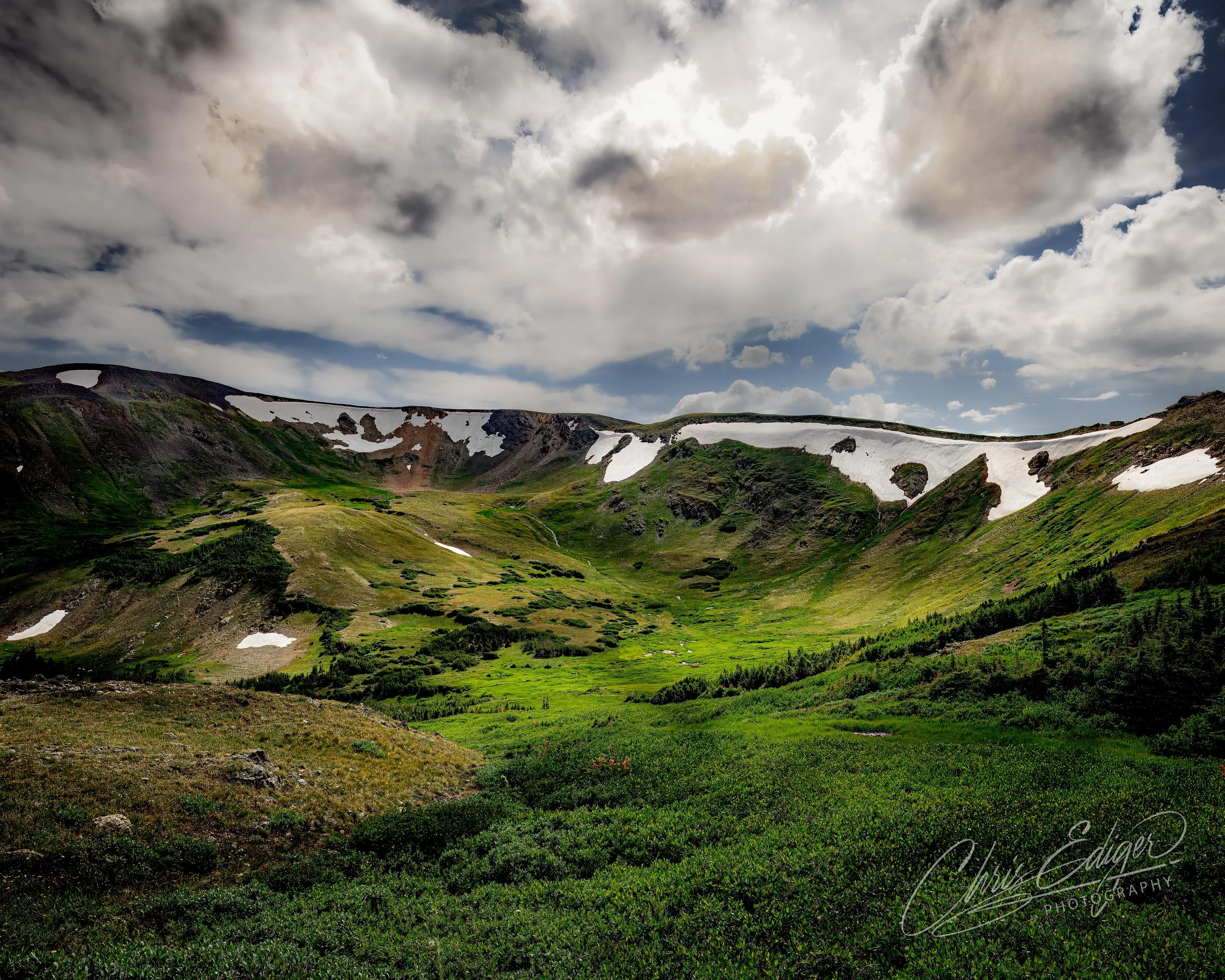 A wide, dramatic view of an alpine basin in summer, surrounded by rugged peaks still clinging to patches of melting snow. The rolling green meadow stretches into the distance, marked by winding game trails and dotted with remnant snowbanks. Above, th