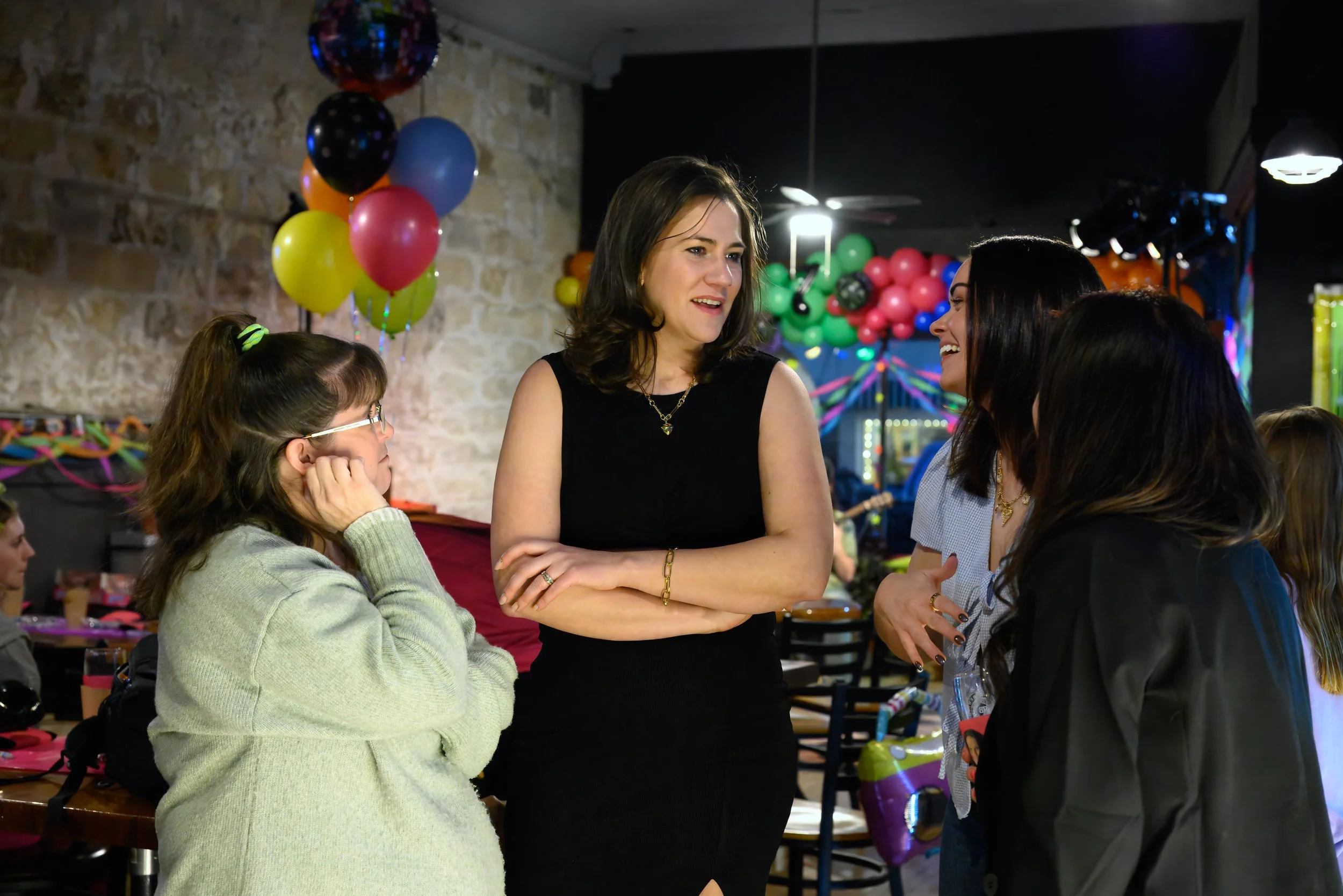 Guests interacting and laughing at a birthday celebration with colorful balloons in the background, photographed as natural candid event coverage.