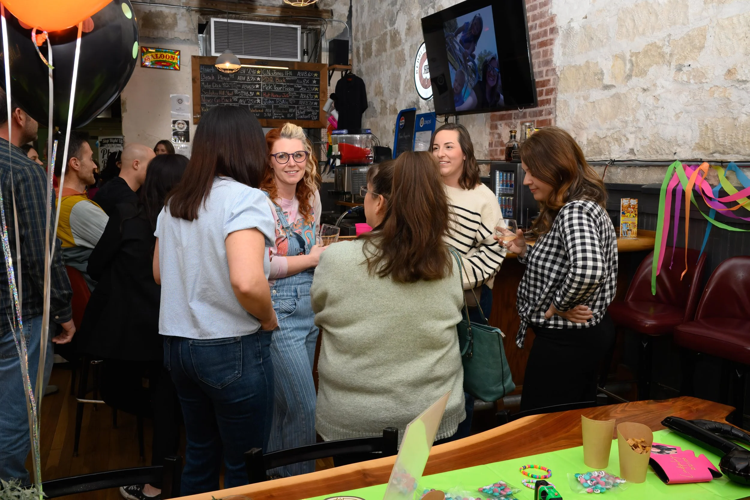 Small group of guests talking and laughing around a bar area during a private event, photographed in a candid documentary style with warm indoor lighting.