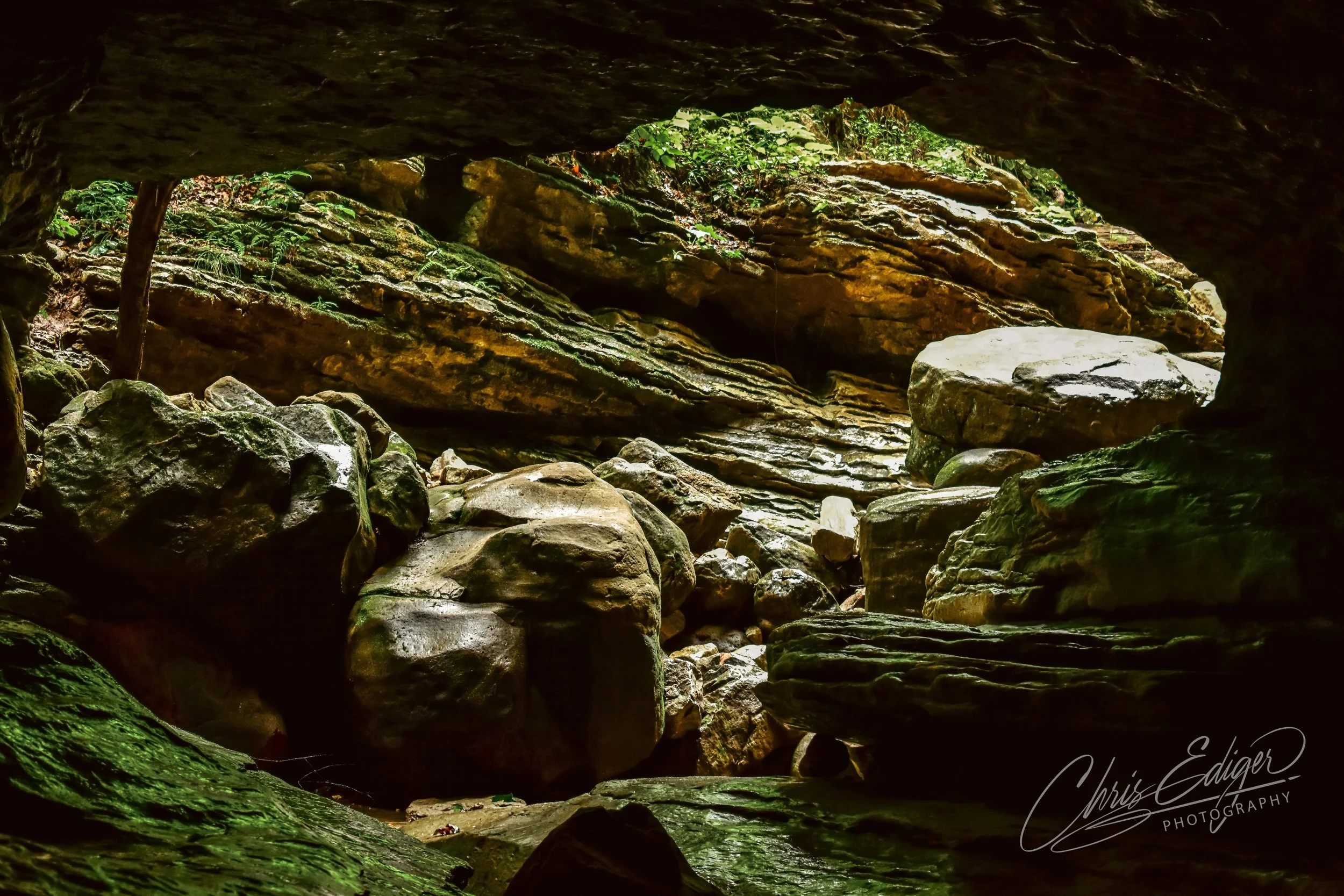 A view from inside a cave looking outward toward a rocky landscape of layered sandstone and moss-covered boulders. Soft natural light filters through the opening, revealing textures of wet stone and green vegetation growing along the rocky walls.