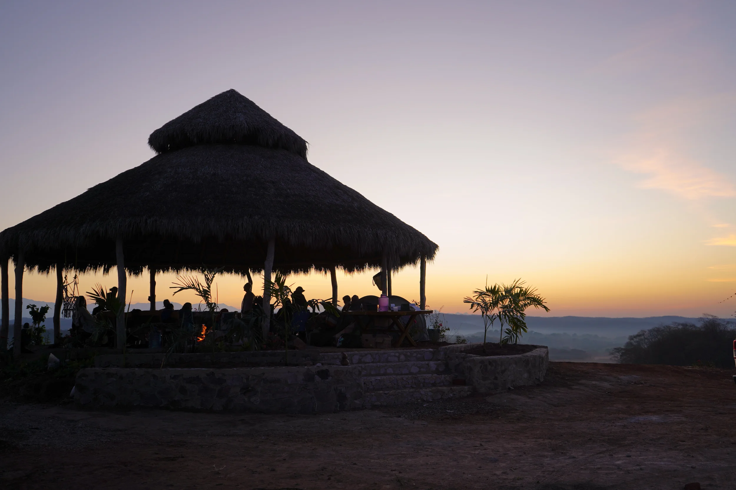 Silhouette of a thatched roof pavilion with people sitting inside, surrounded by plants, during a sunset with a hazy landscape in the background.