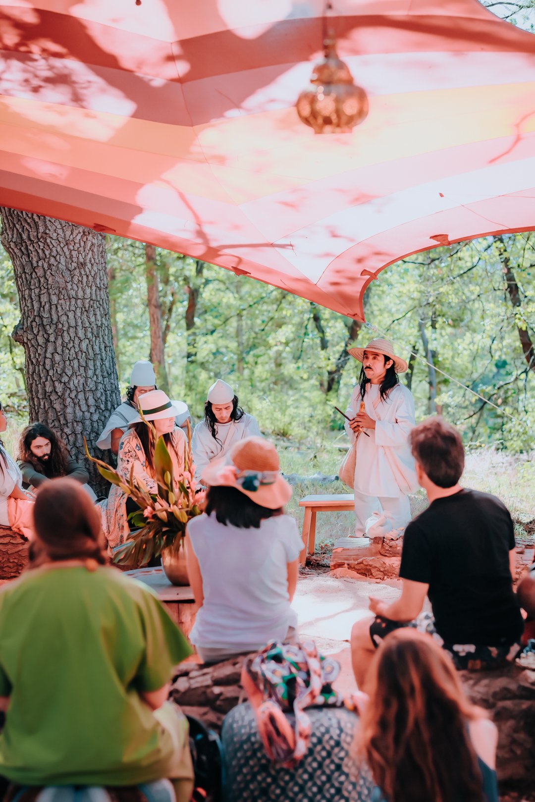 People gathered outdoors in a forest, sitting under a large pink canopy. A woman in white with a straw hat appears to be leading a discussion or presentation, surrounded by others seated on logs.