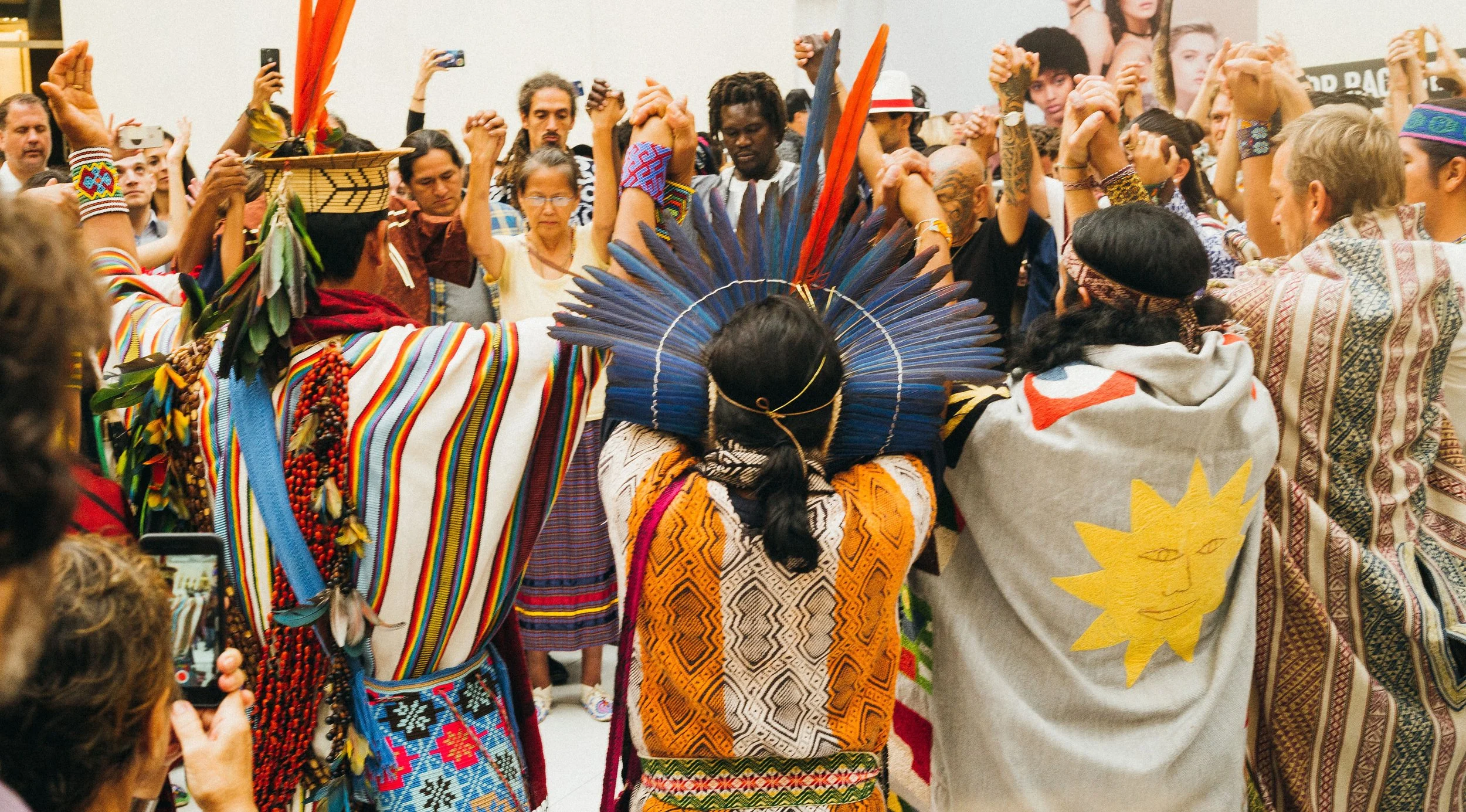 Indigenous people and spectators participating in a cultural ceremony or celebration, wearing traditional clothing and headdresses, holding hands in a circle.