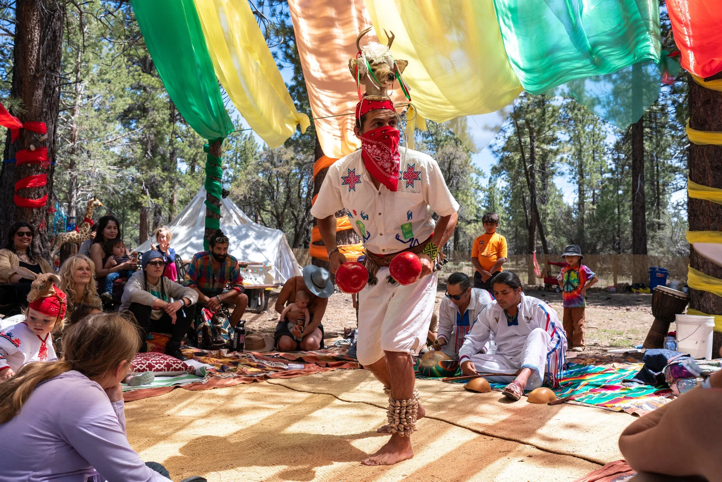 A performer in colorful attire with face paint and shell ankle cuffs juggling red balls during an outdoor event surrounded by an audience.