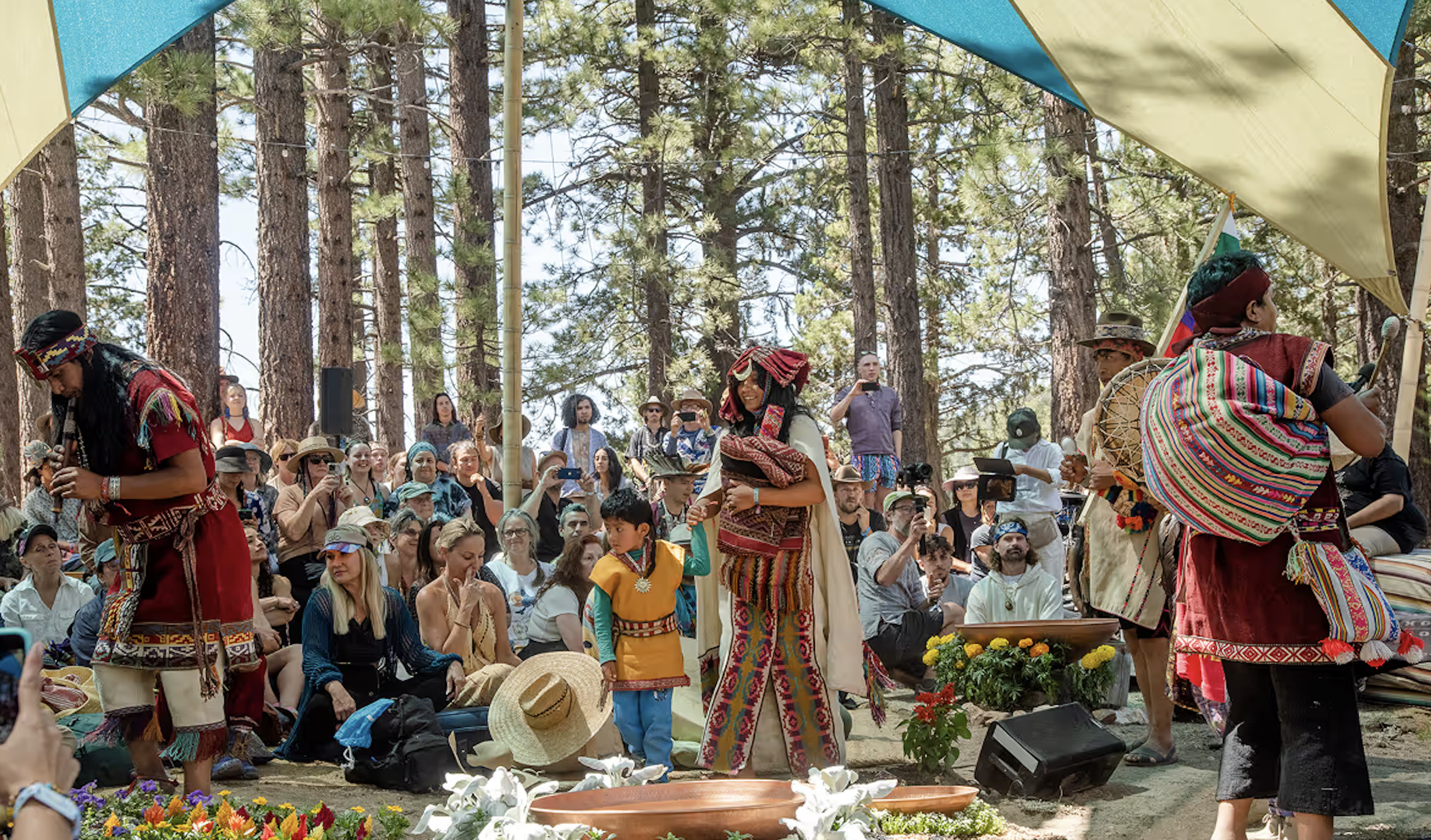 People gathered outdoors among tall trees, participating in a cultural event wearing traditional colorful clothing and accessories, with some holding objects and smiling.