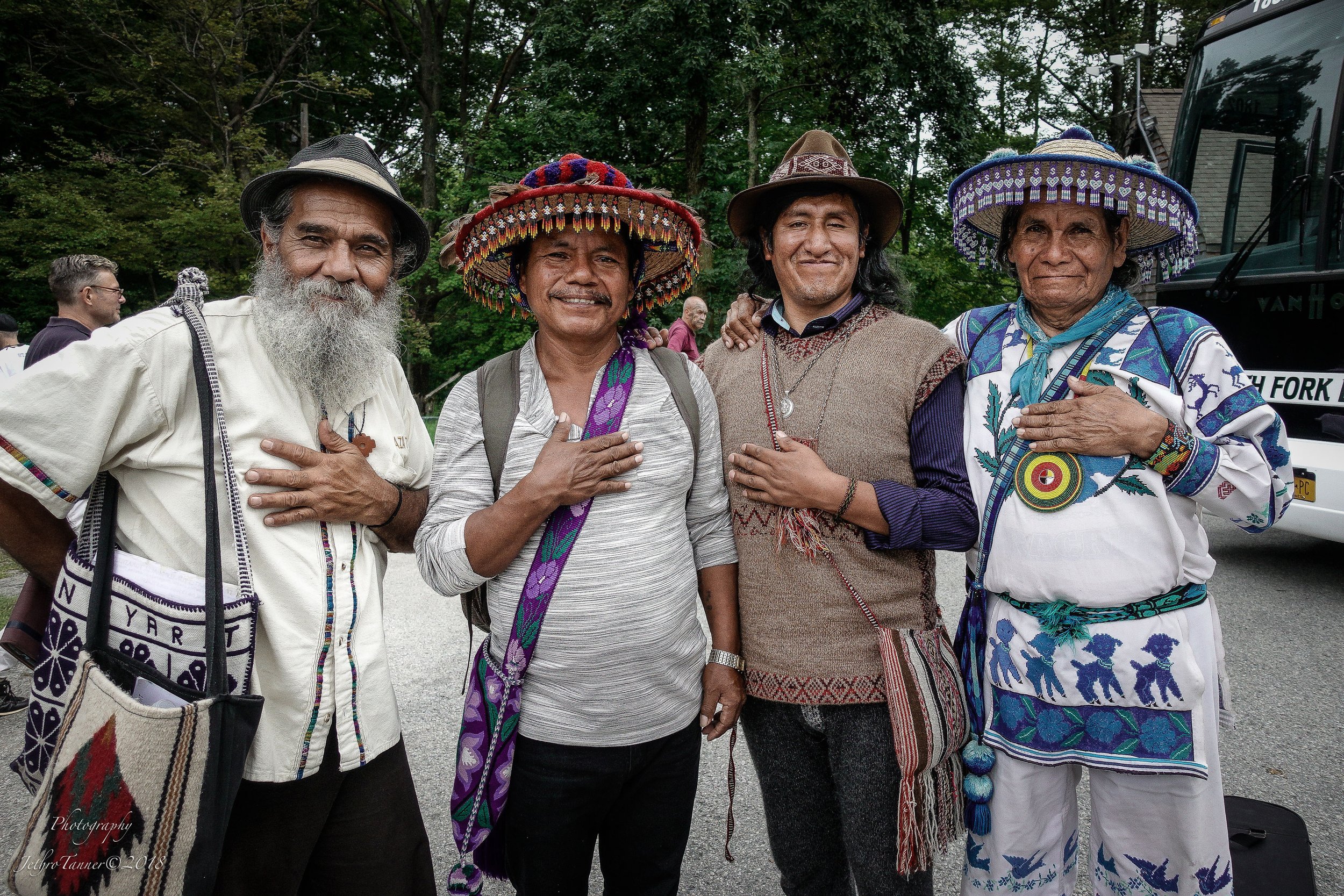 Four men standing outdoors with trees in the background, wearing traditional Indigenous clothing and hats, with some placing their hand over their heart.