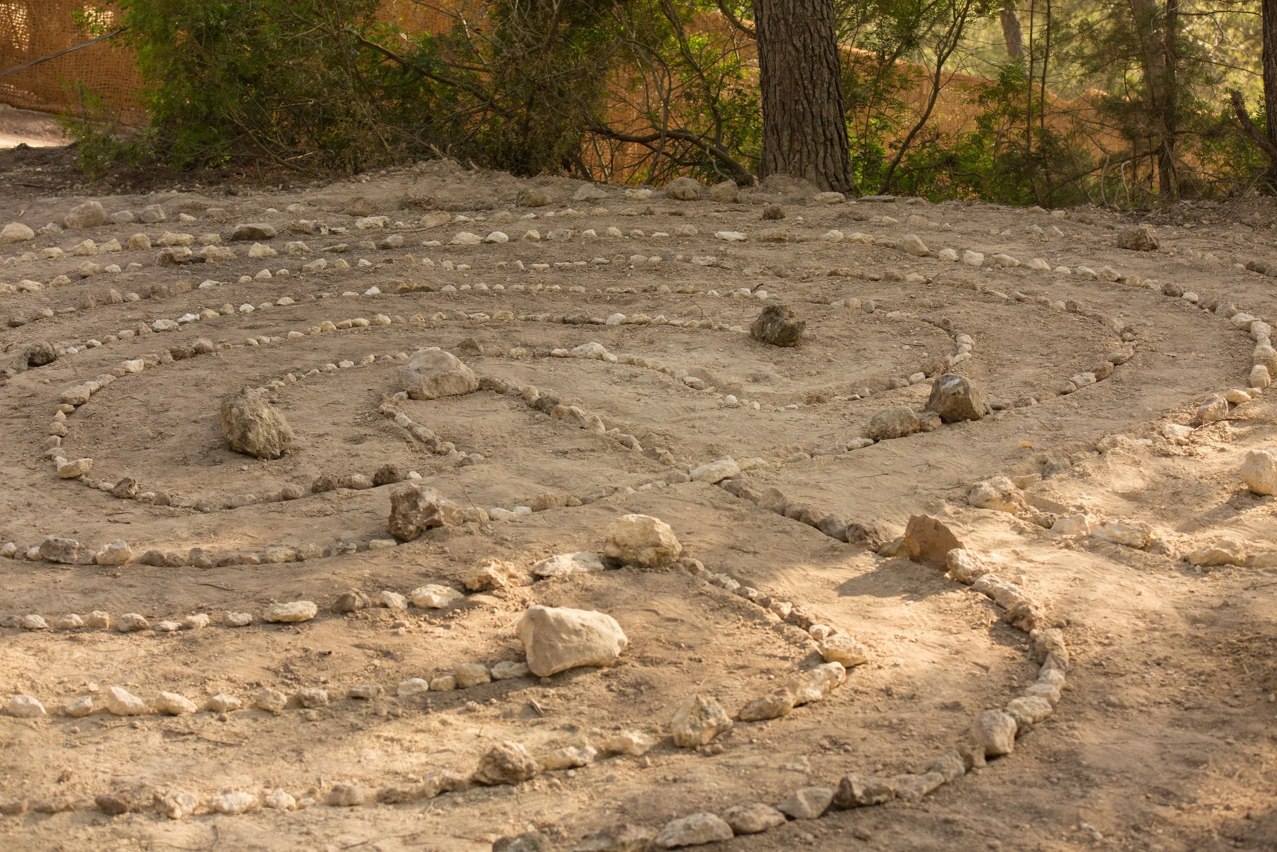 A dirt ground with a spiral pattern made of small stones, surrounded by natural trees and bushes.